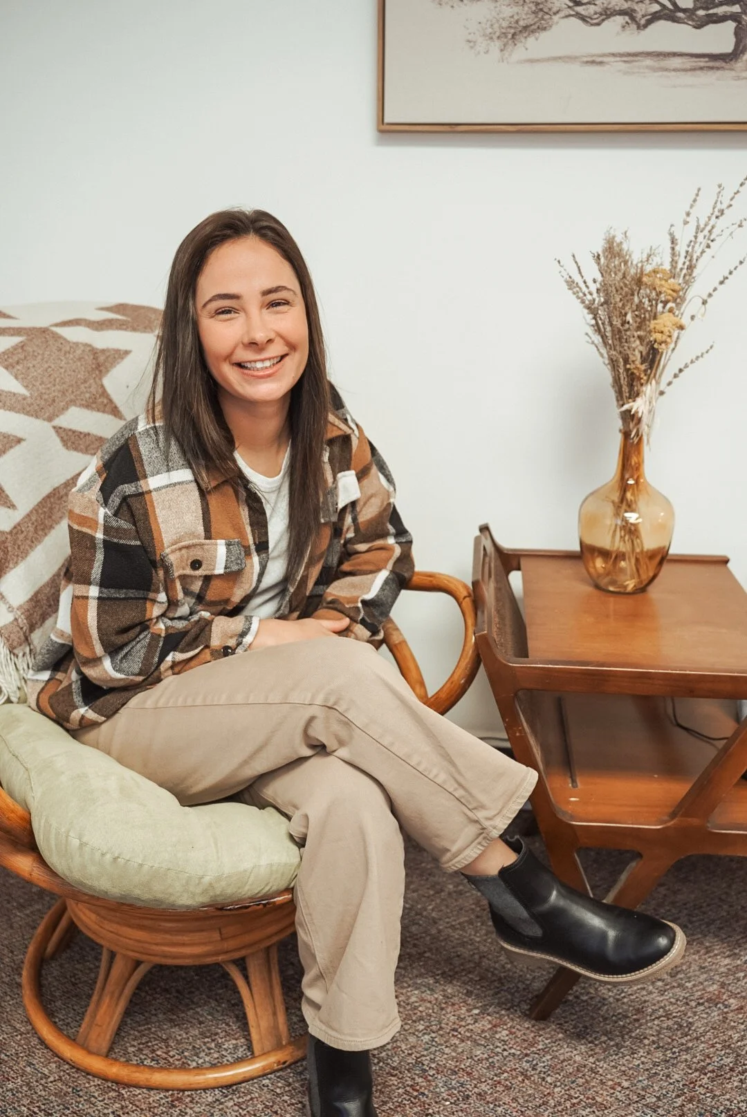 A woman sitting on a cushioned wooden chair, smiling, wearing a plaid jacket, beige pants, and black boots, with a wooden side table and a vase of dried flowers in the background.
