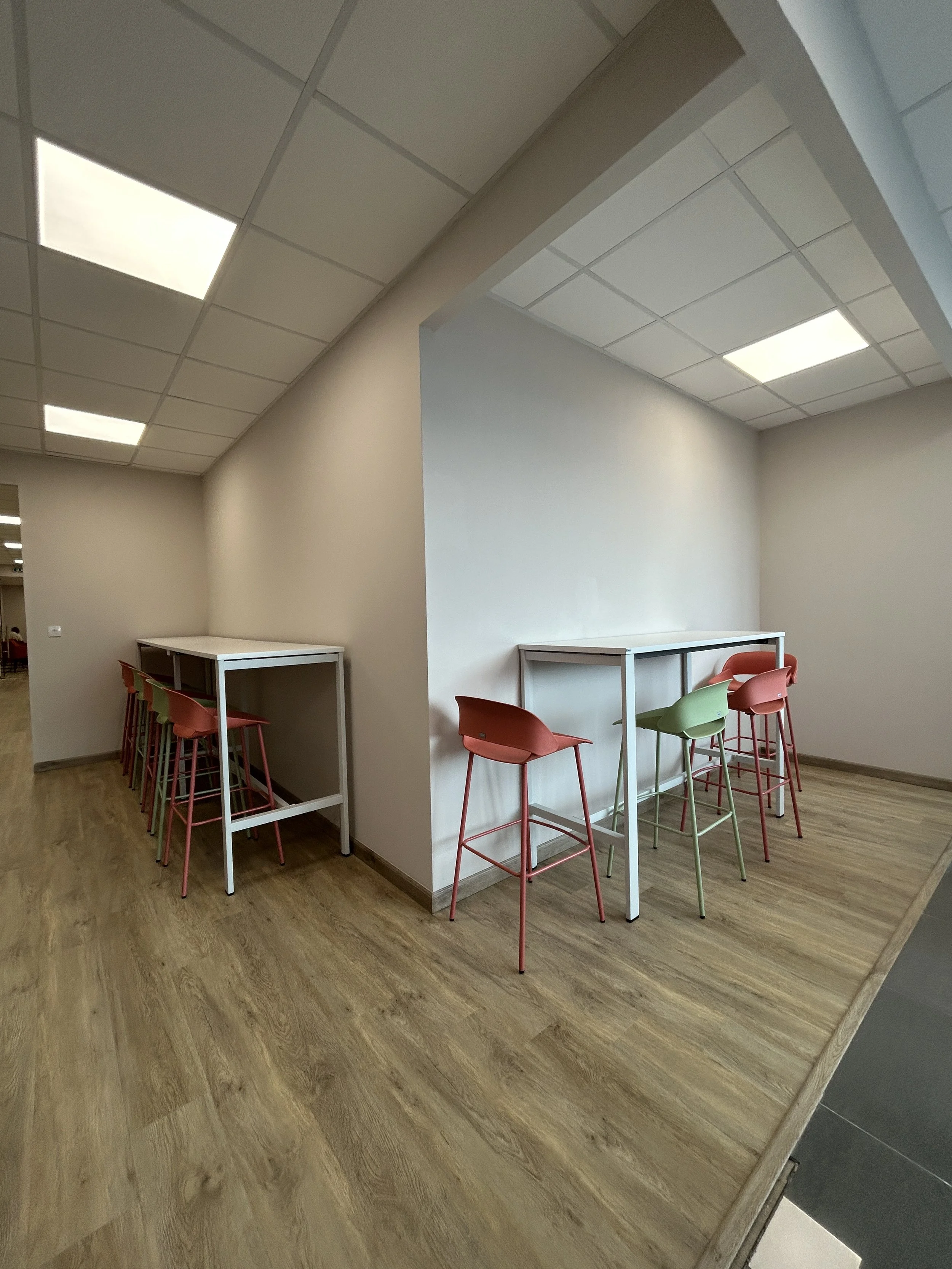 Empty seating area with colorful high chairs and white tables in a modern room with wooden floors and a ceiling with fluorescent lights.