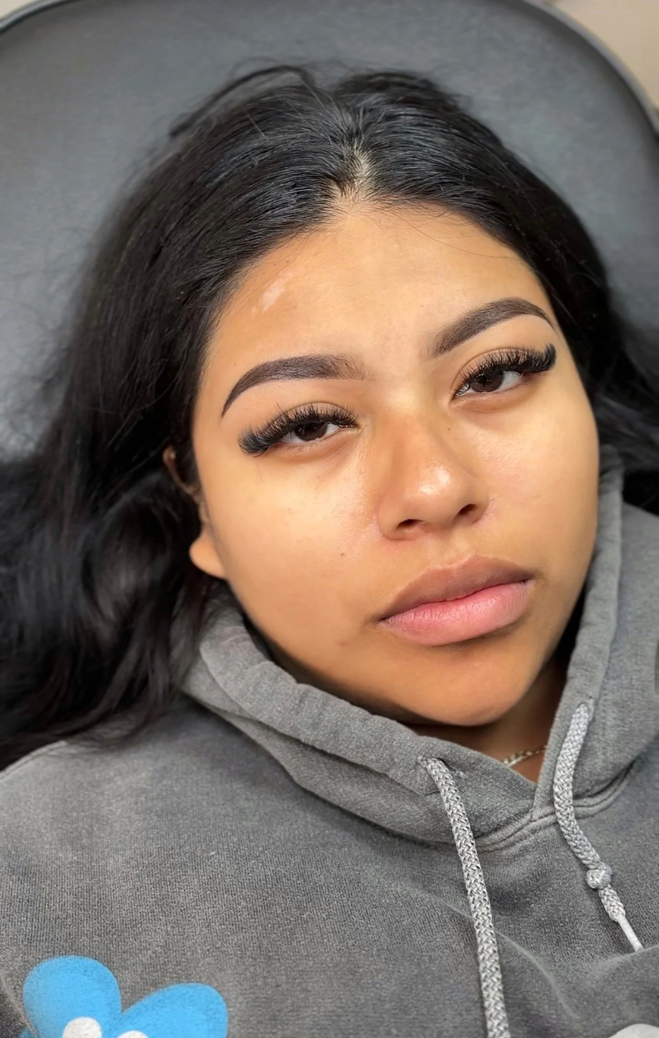 Close-up of a young woman with long black hair, wearing a gray hoodie, lying down on a gray surface, looking at the camera.