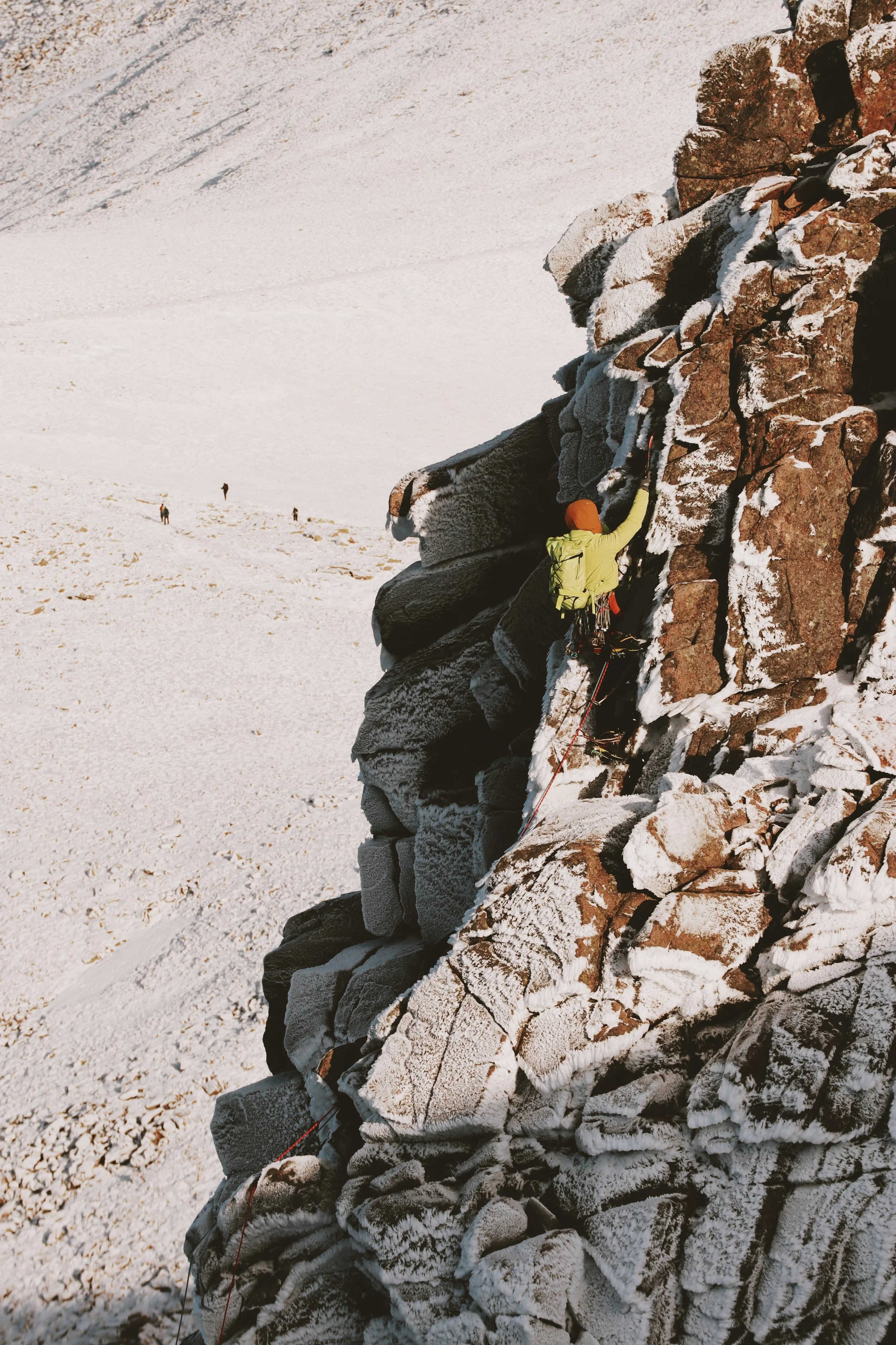 A person wearing a yellow jacket, orange beanie, and climbing gear ascends a snow-covered rock face in a mountainous landscape with snowy slopes and other climbers in the background.