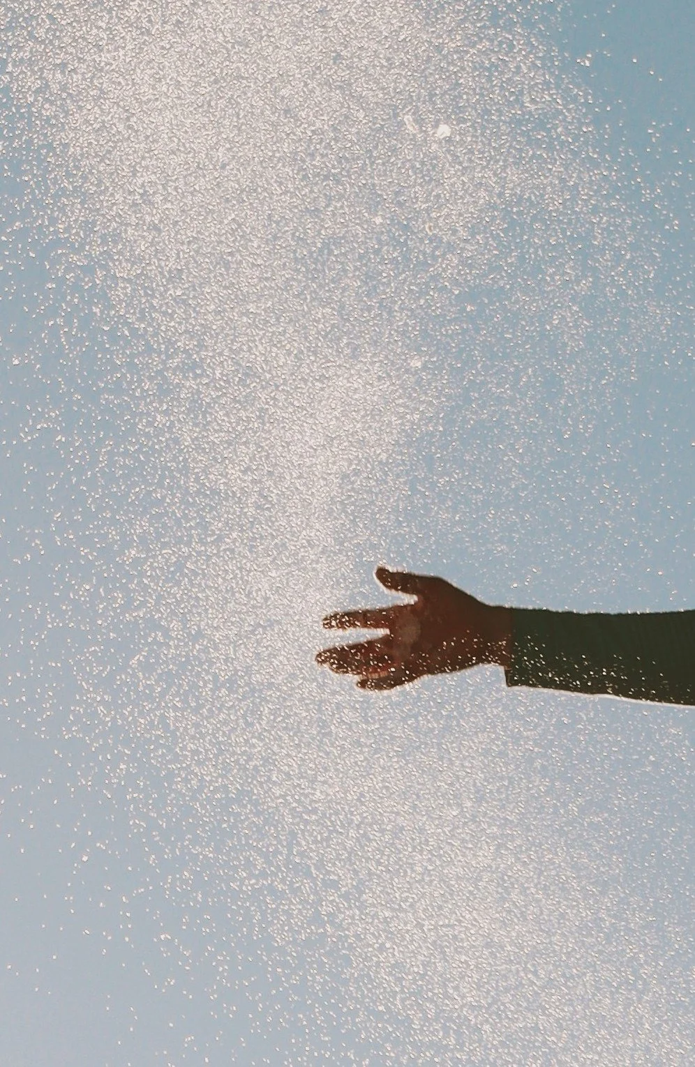 A person's hand reaching out towards a light blue sky with tiny sparkling particles, possibly snow or glitter, scattered across the scene.