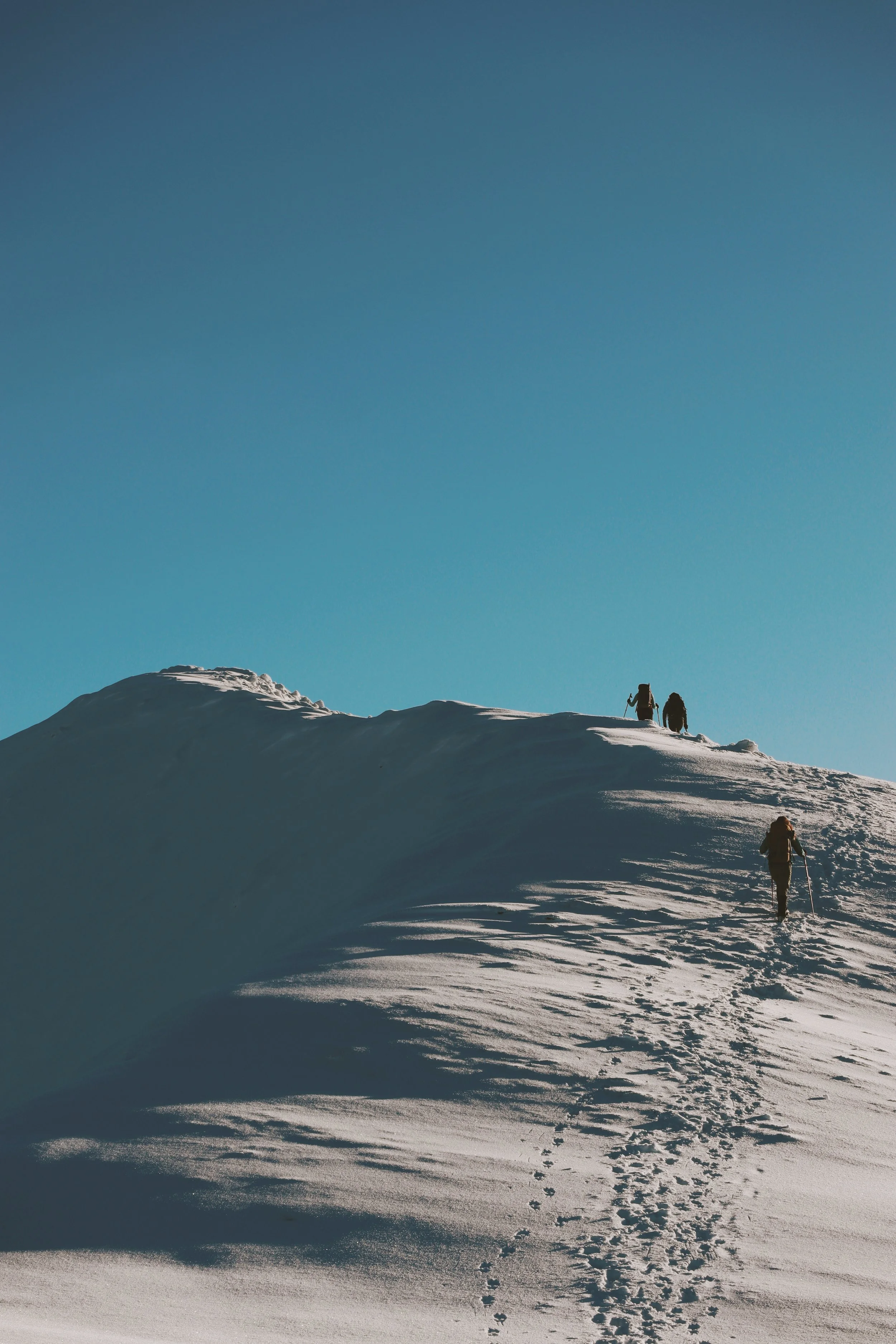 Three people hiking up a snow-covered mountain under a clear blue sky.
