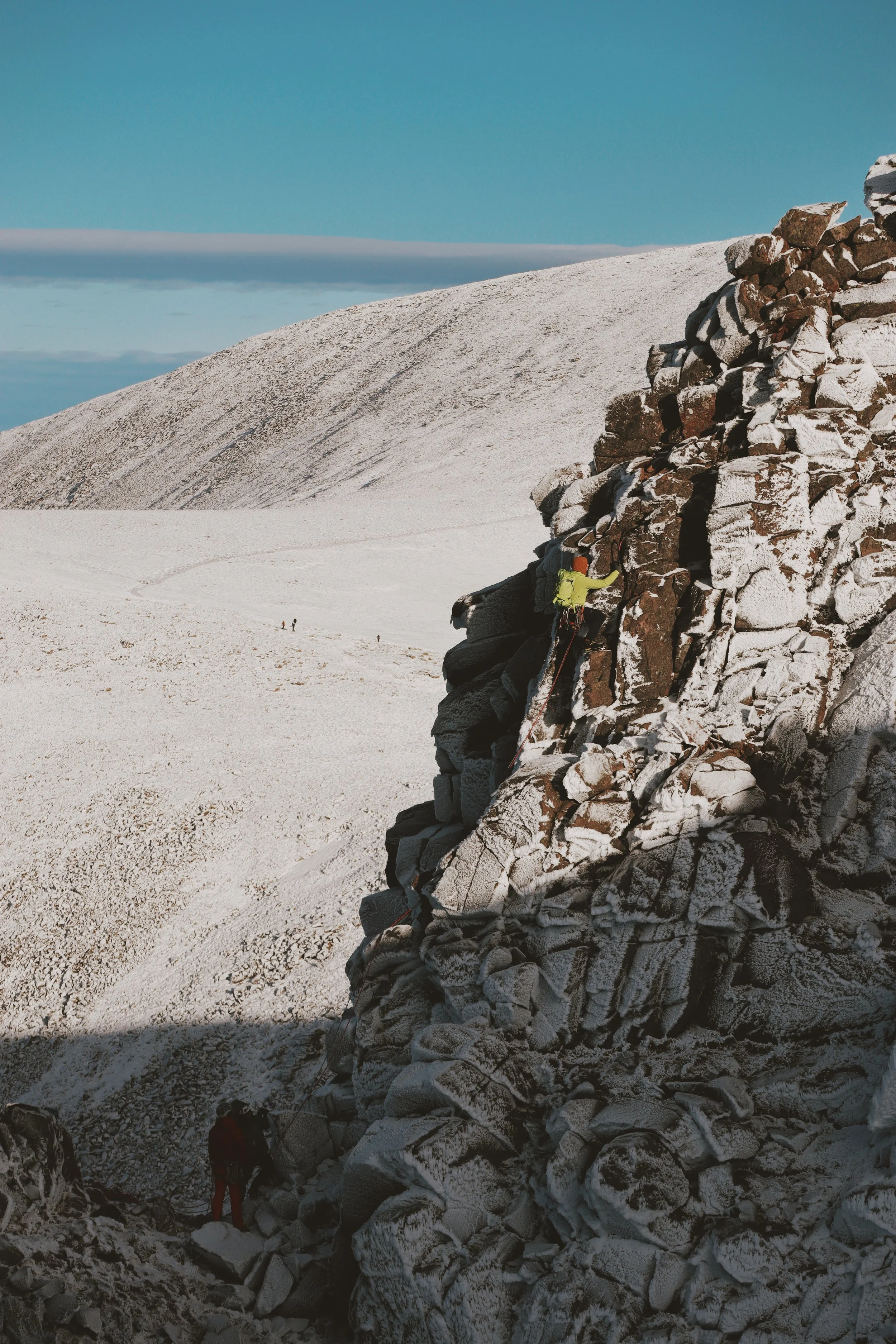 A person climbing a rocky, snow-covered mountain face with safety equipment, set against a snowy landscape and clear blue sky.