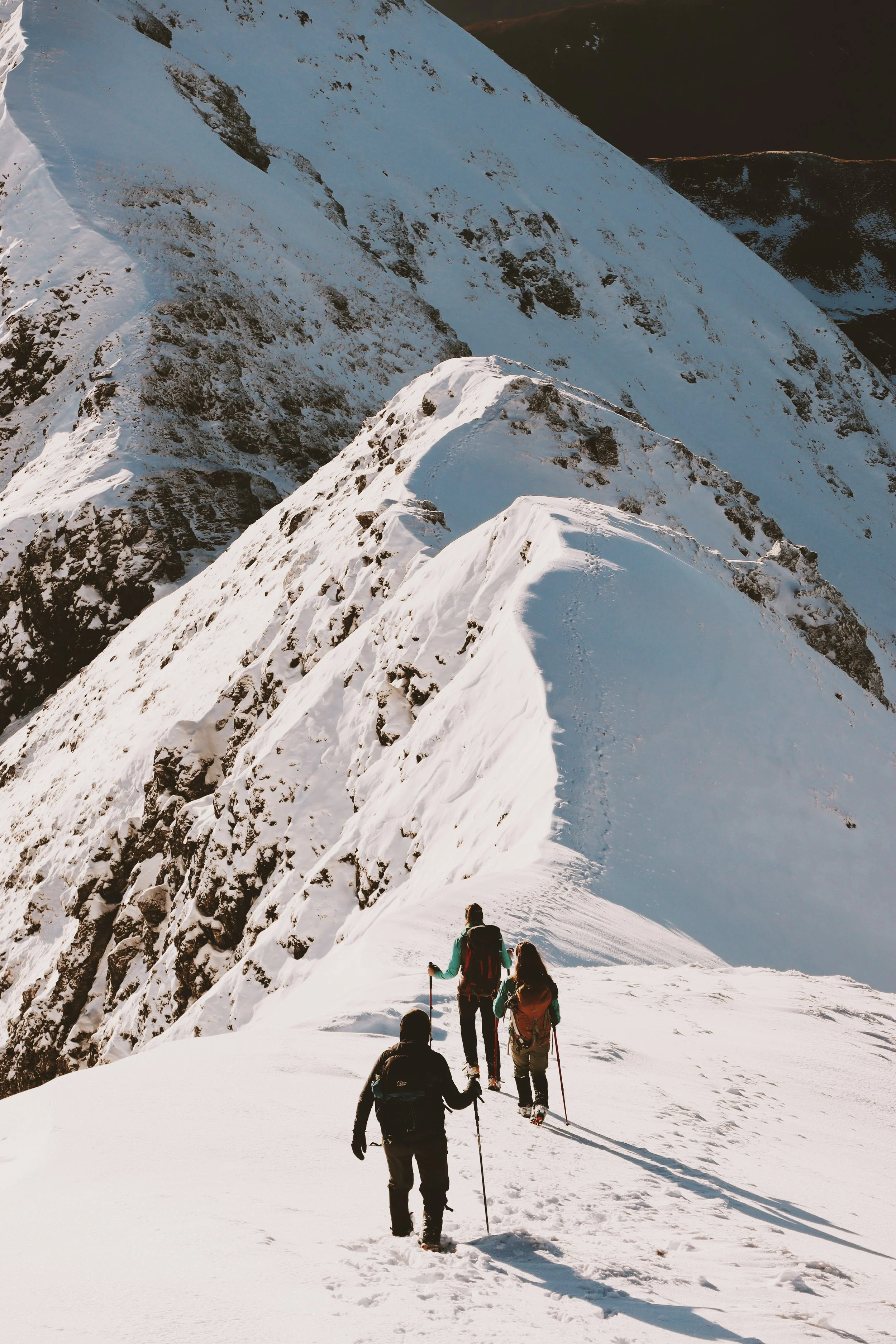 Four hikers ascending a snow-covered mountain trail with rocky peaks in the background.