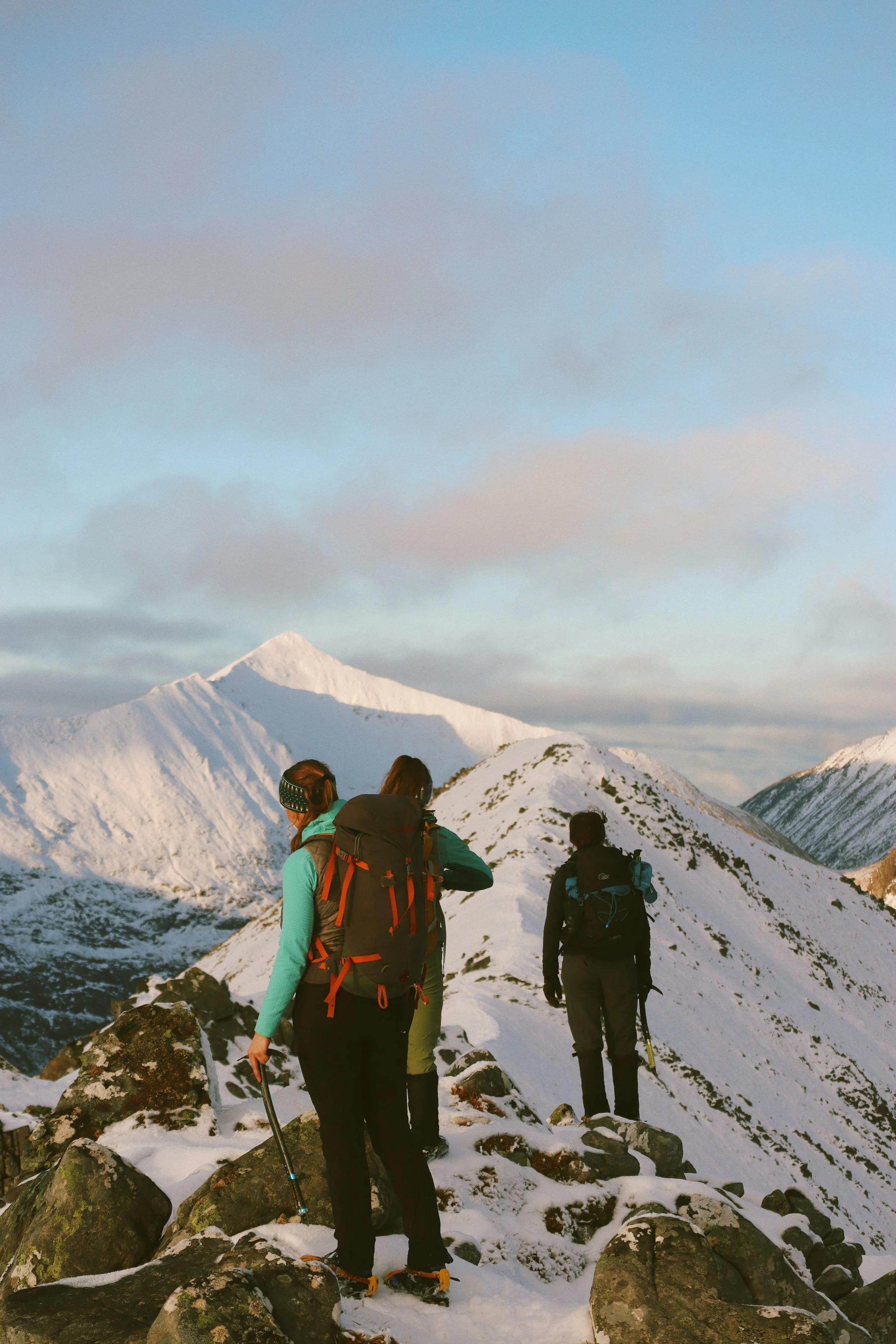 Three hikers with backpacks and hiking gear standing on a snow-covered mountain trail, looking at snow-capped peaks and a partly cloudy sky.