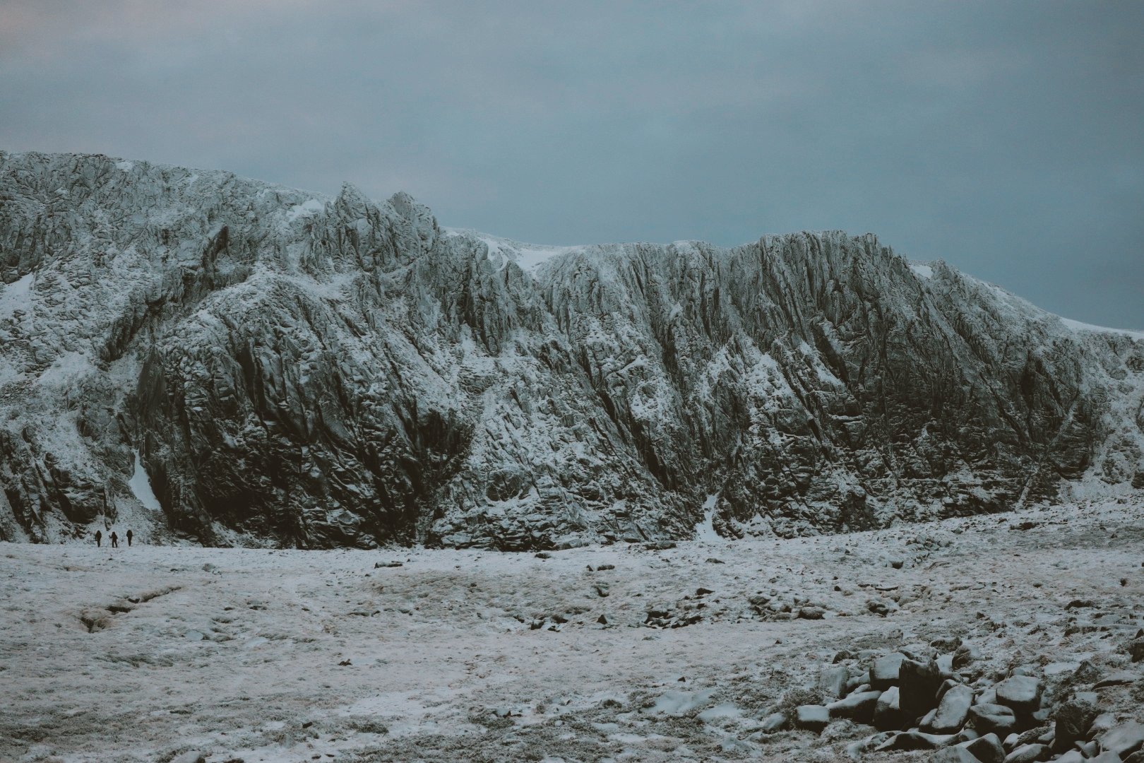 Snow-covered mountainous landscape with three tiny figures walking in the distance and rocks in the foreground.