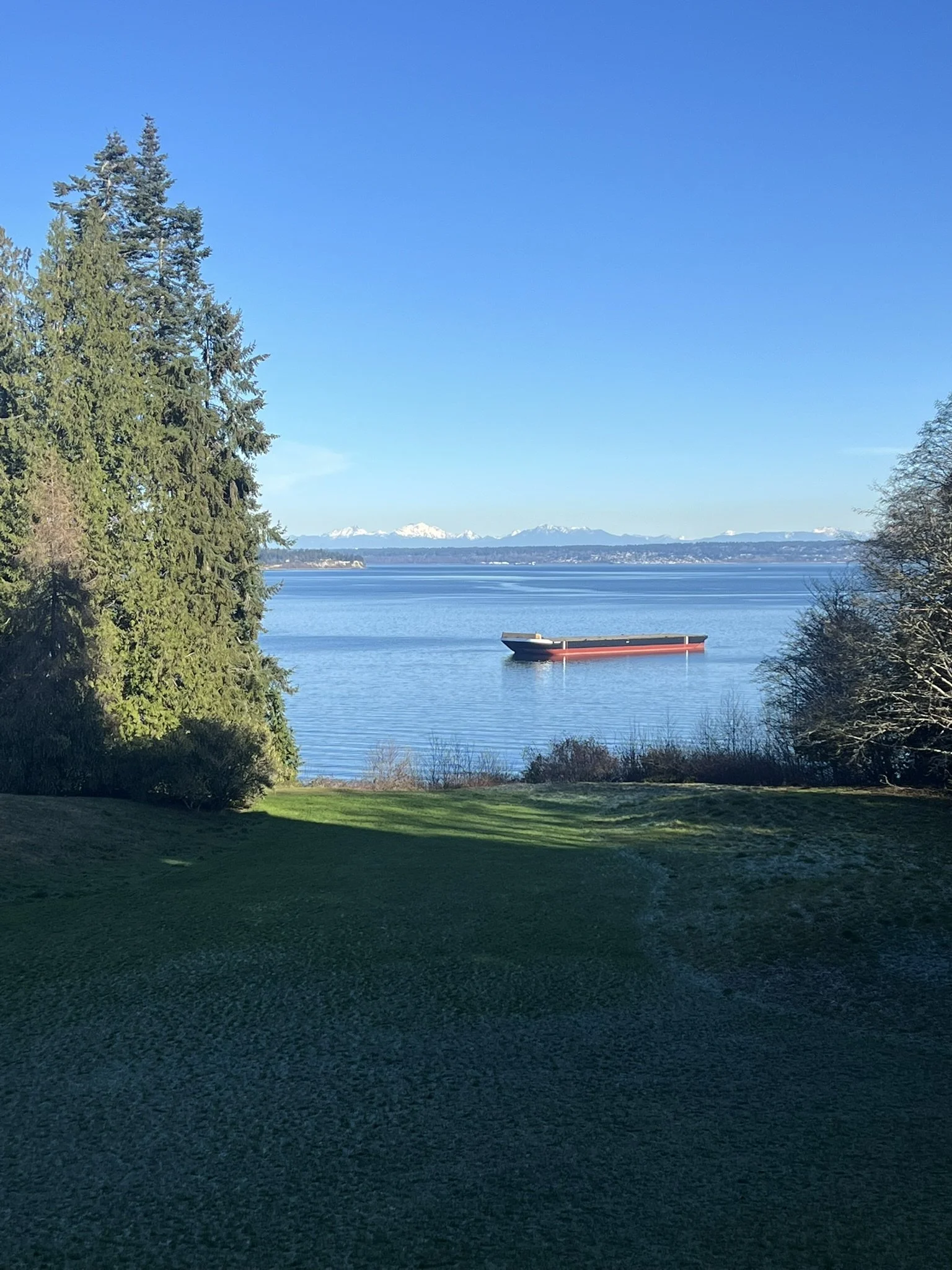  A barge floats on calm blue waters off Bainbridge Island, framed by evergreen trees and a wide grassy lawn at Bloedel Reserve, with snow-capped Cascade Mountains visible in the distance. 