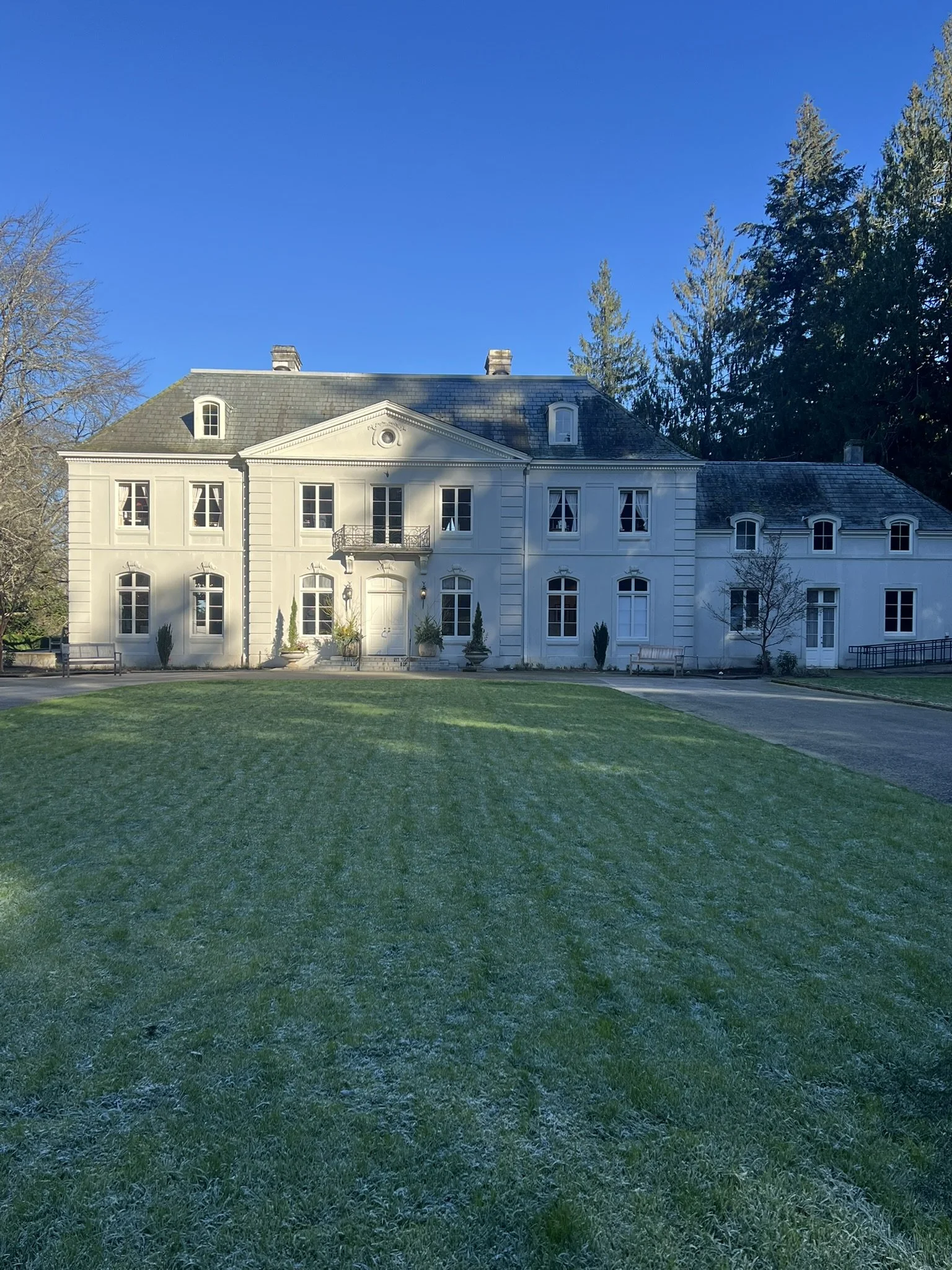  The historic Bloedel Residence, a stately mansion with dormer windows and a slate roof, sits beneath a clear blue sky, framed by evergreens and a wide manicured lawn. 