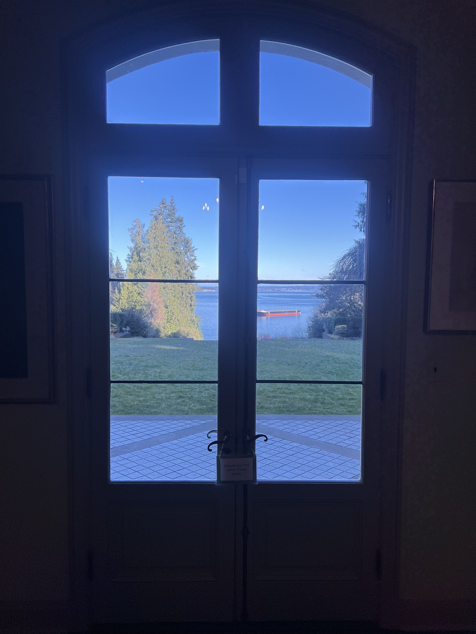  View through a tall arched window at Bloedel Reserve Residence, looking out onto a lawn, tiled terrace, and Puget Sound with a red barge in the distance under a clear blue sky. 