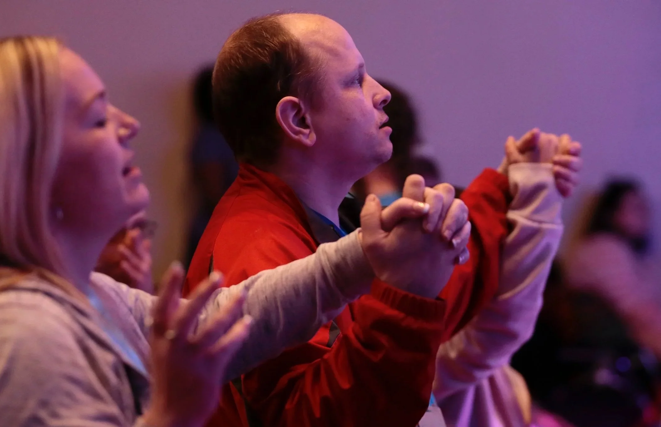 Two adults hold hands and raise their arms in worship during a church service, standing among others in a softly lit, sensory-friendly environment.