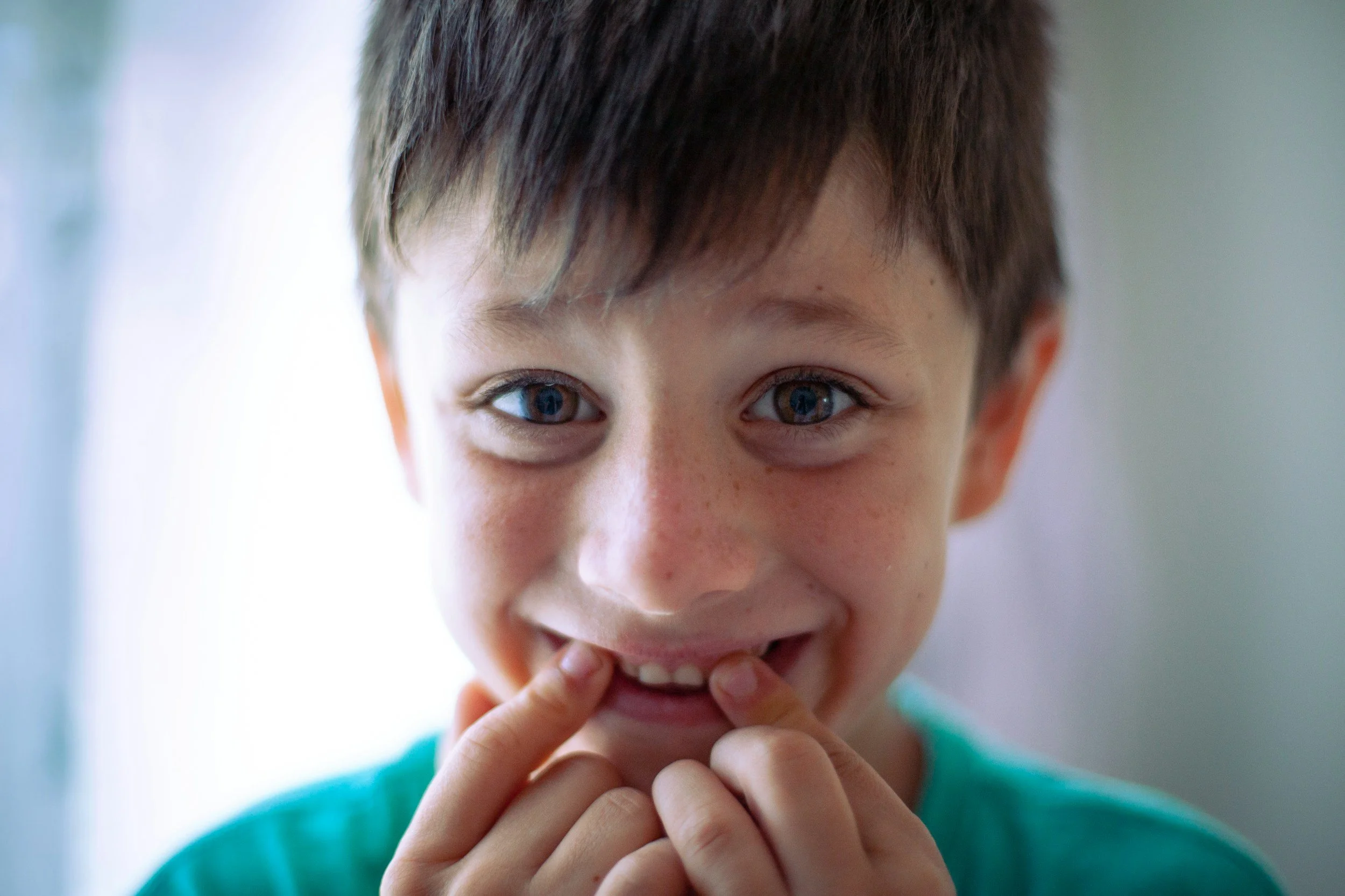 Close-up of a young child with brown hair and bright eyes, smiling with fingers near his mouth, wearing a teal shirt.