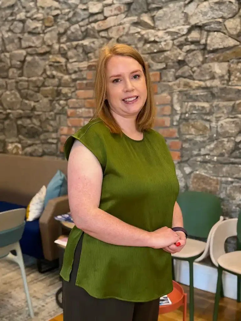 A woman with red hair wearing a green top and black pants standing indoors against a stone wall with chairs and a sofa in the background.