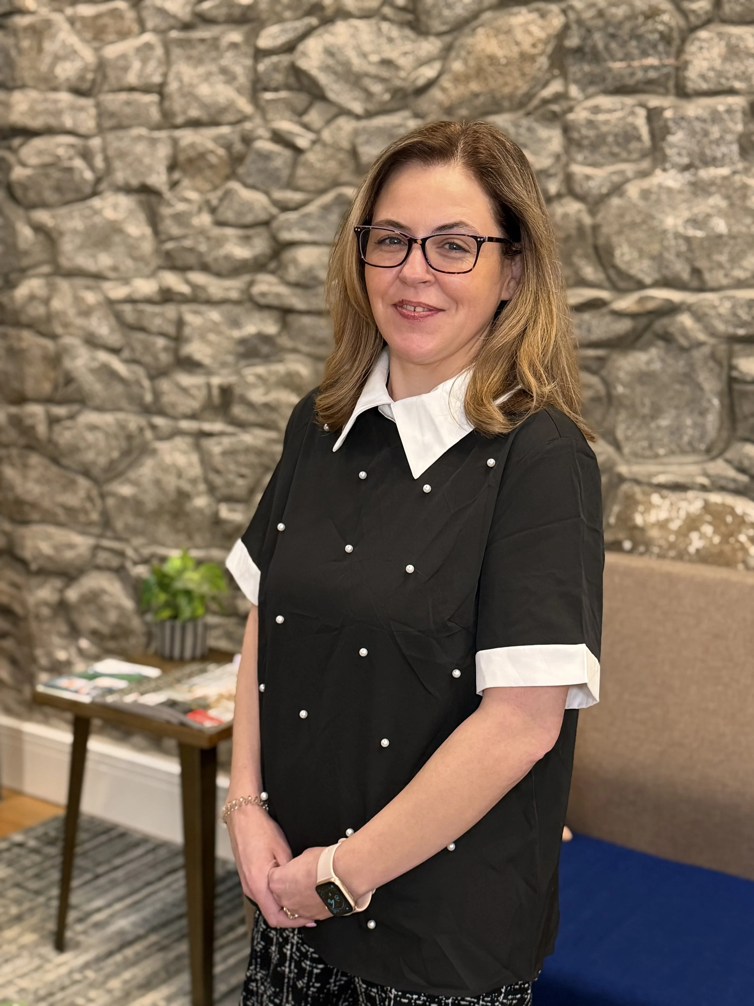A woman with shoulder-length brown hair, glasses, and a white collared shirt under a black dress with pearl-like buttons, standing in front of a stone wall. She is smiling and has her arms crossed.