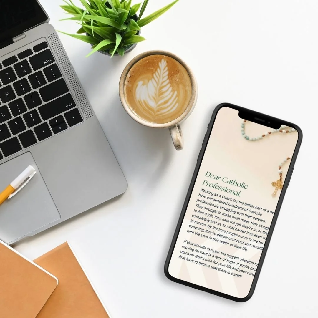 A flat lay of a workspace with a laptop, a smartphone displaying a message reading "Dear Catholic professionals...", a cup of latte with latte art, a small potted plant, two brown notebooks, and a pen on a white desk.