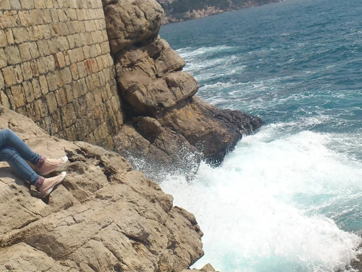 Rocky seaside cliff with a stone wall, pink shoes, and waves crashing below