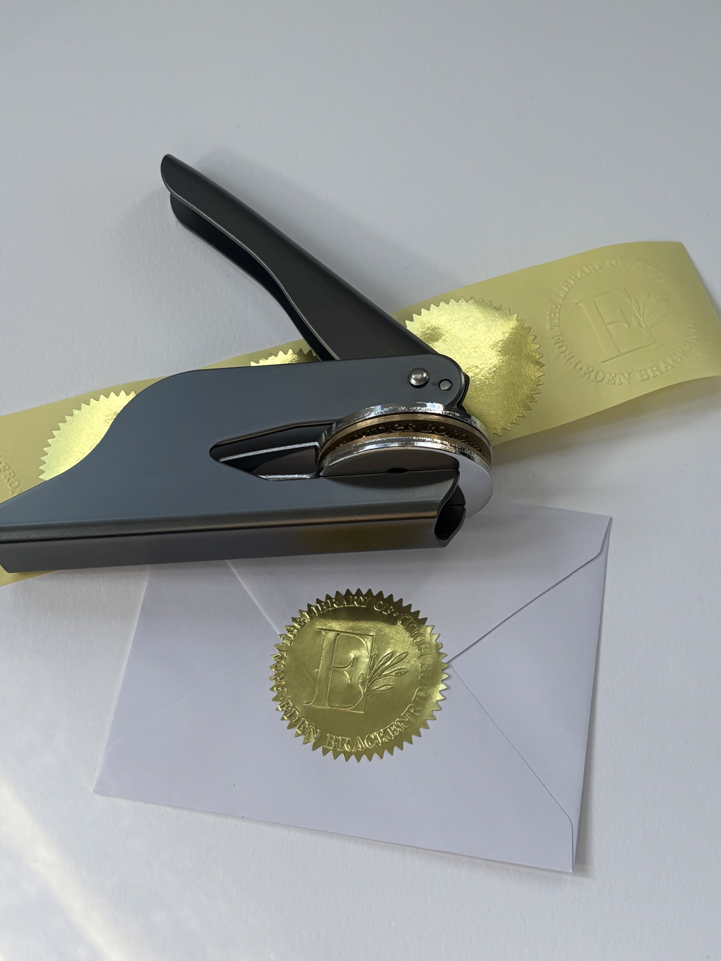 Gunmetal embosser laying on top of blank gold foil stickers. Pressed gold foil sticker used to seal envelope.