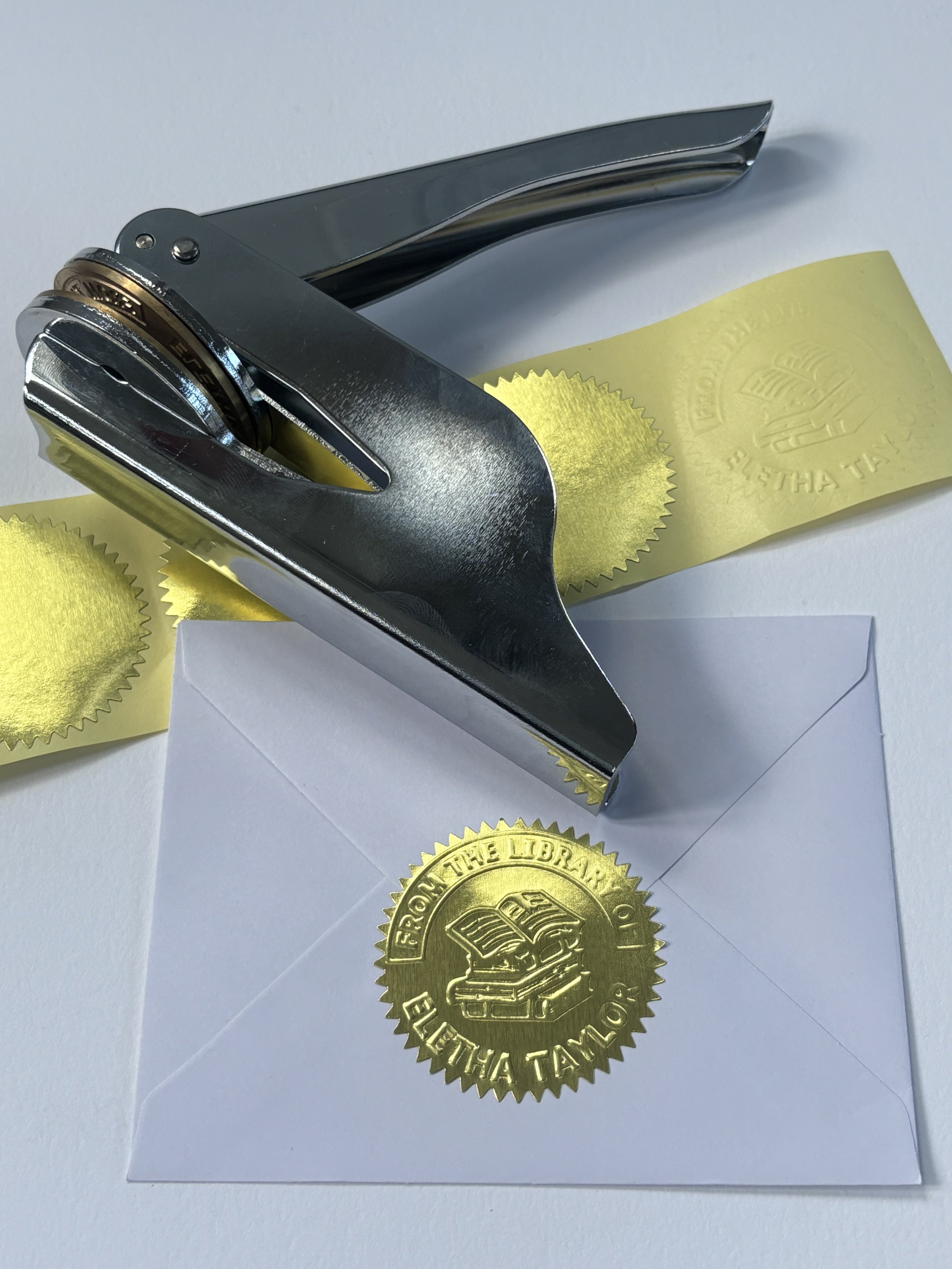 Silver embosser laying on top of blank gold foil stickers. Pressed gold foil sticker used to seal envelope.