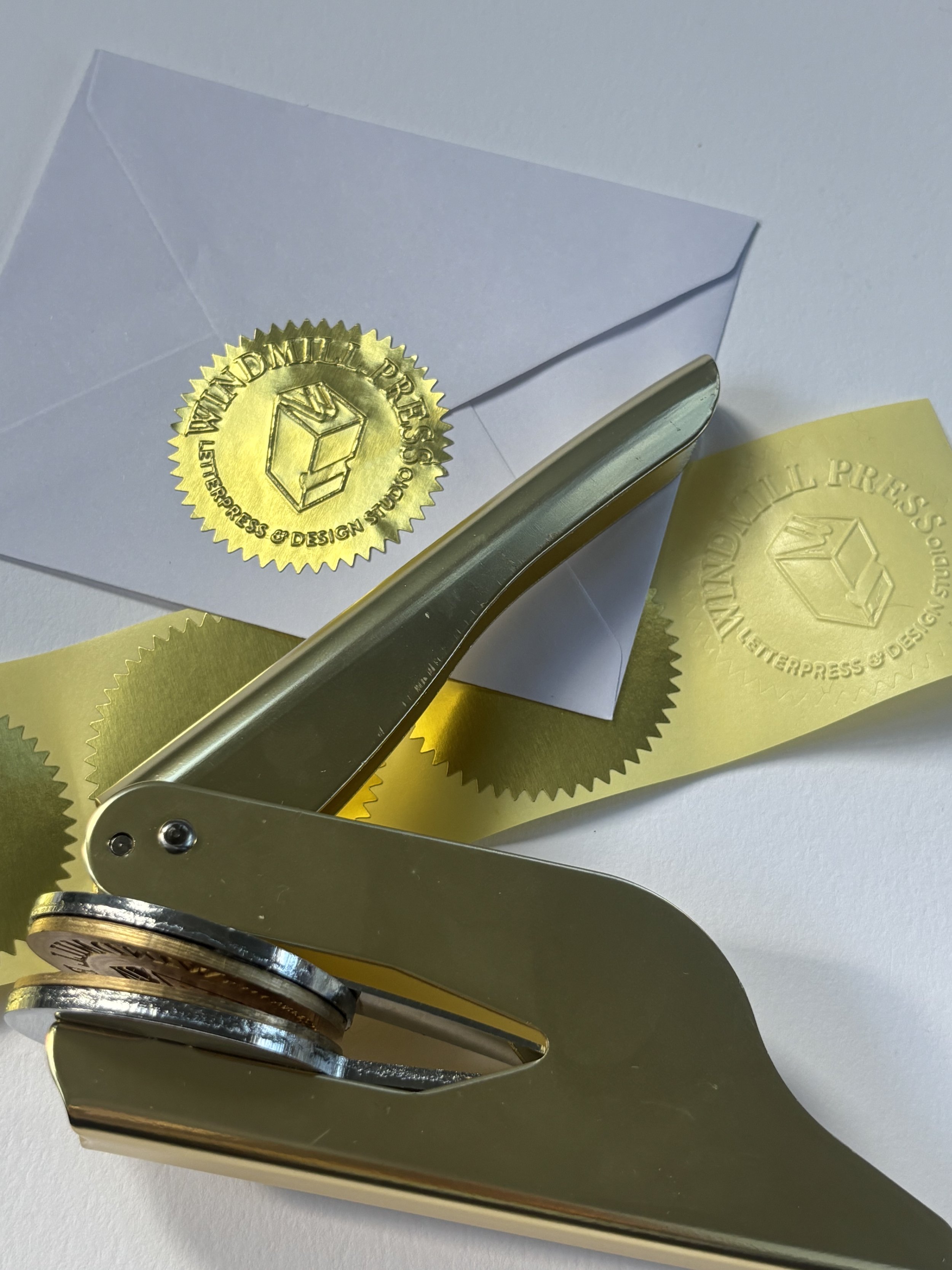Gold embosser laying on top of blank gold foil stickers. Pressed gold foil sticker used to seal envelope.