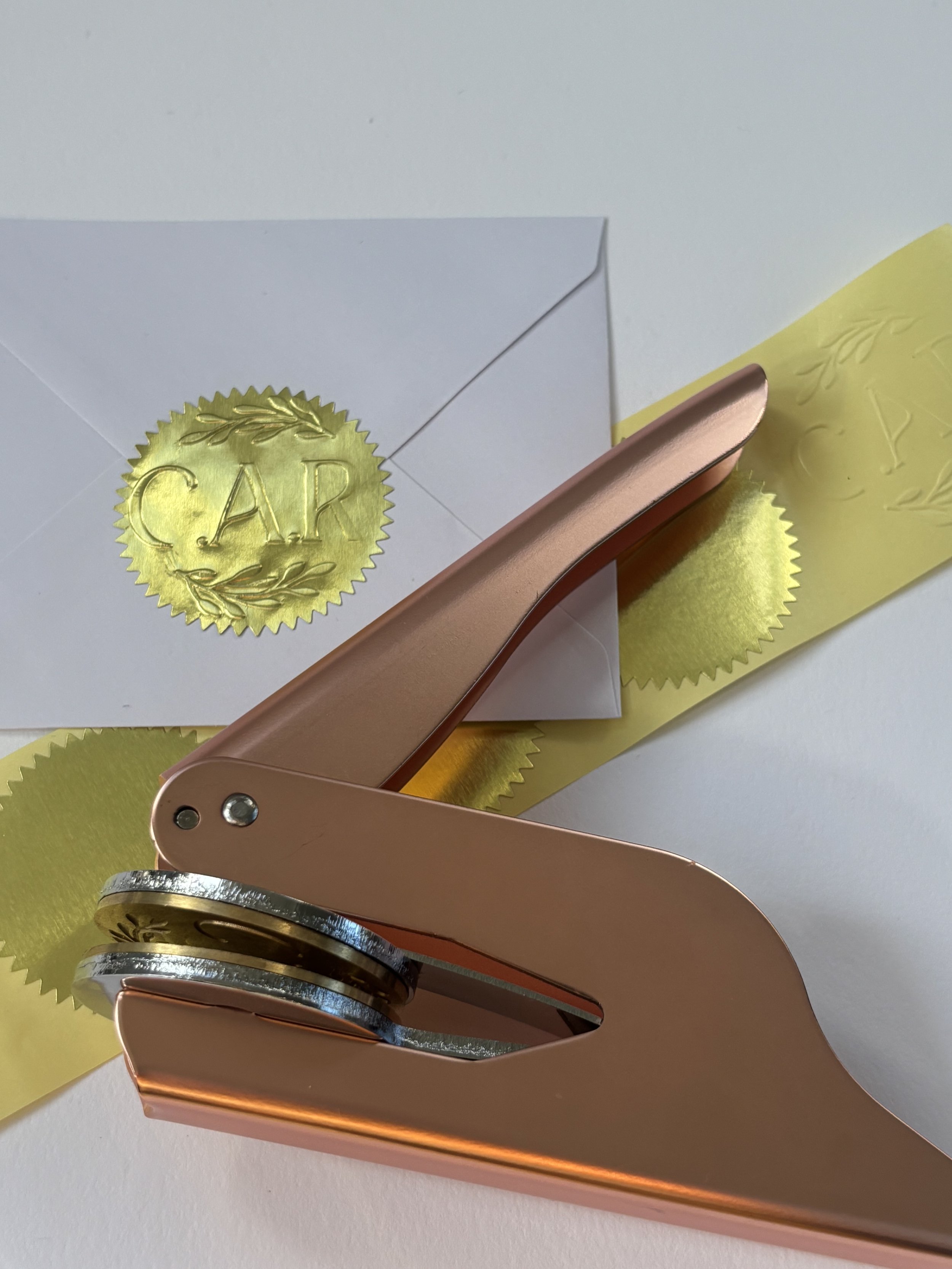Rose gold embosser laying on top of blank gold foil stickers. Pressed gold foil sticker used to seal envelope.