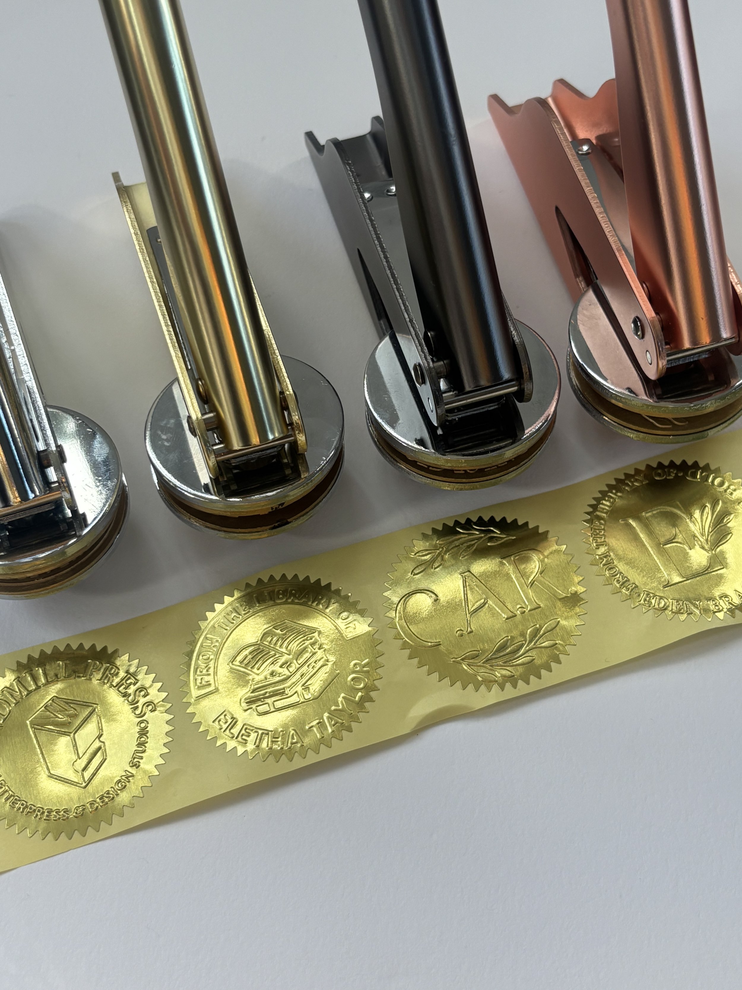Embossers in various metals lined up in front of pressed gold foil stickers. Various designs on stickers.
