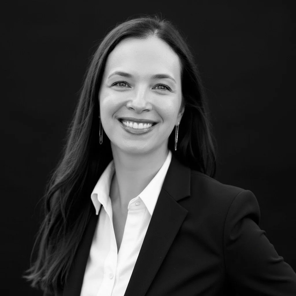 Black and white portrait of a woman with long dark hair, wearing a blazer and a white shirt, smiling against a dark background.