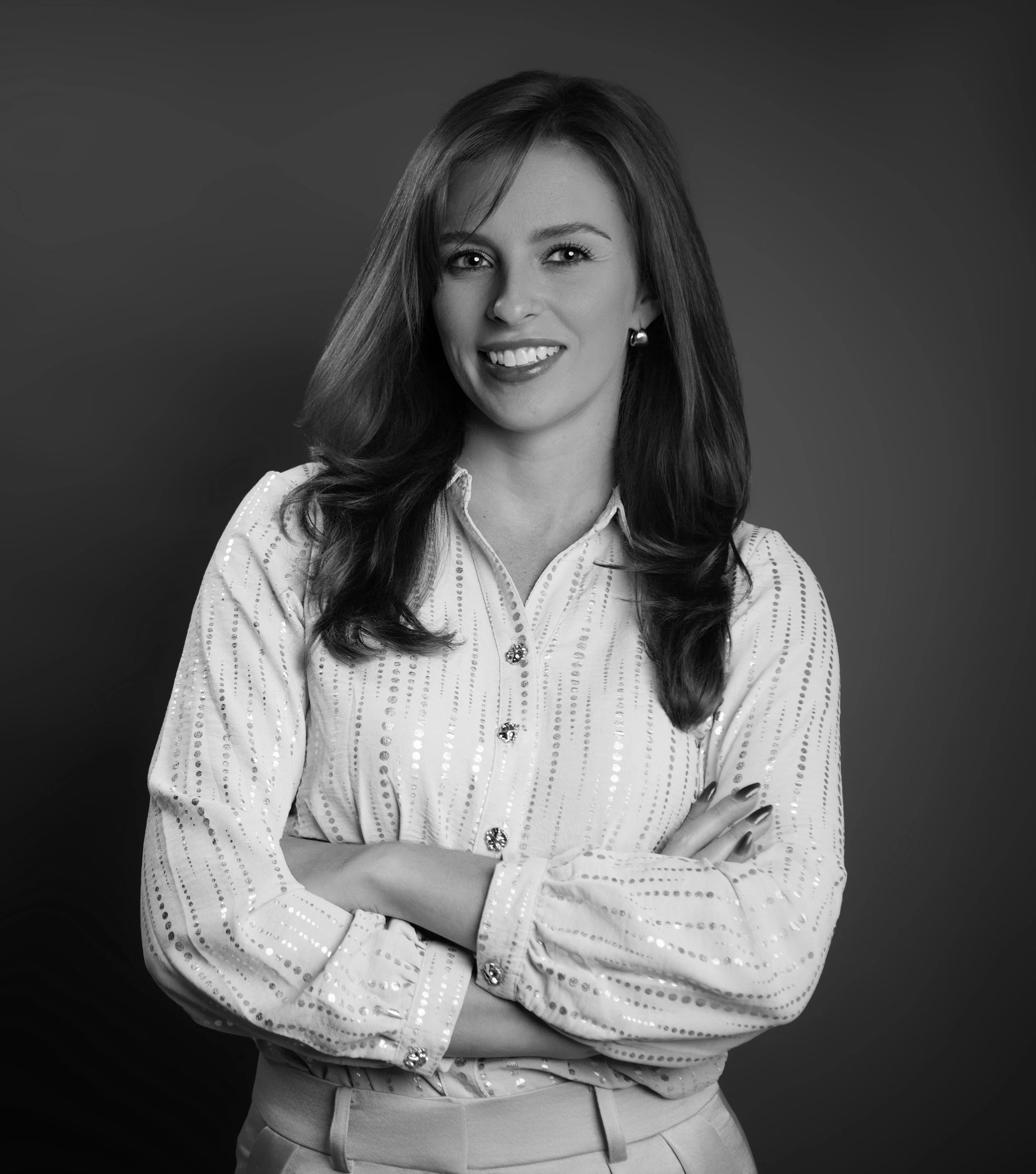 Black and white portrait of a smiling woman with long dark hair, wearing a patterned blouse with rolled sleeves, standing with arms crossed against a plain background.