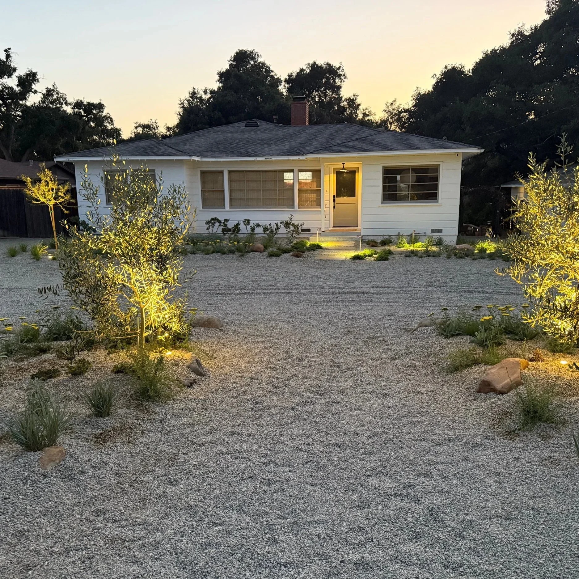 A small white house with a dark roof, lit from outside at dusk, with a gravel front yard landscaped with young bushes and decorative rocks, illuminated by outdoor lighting.