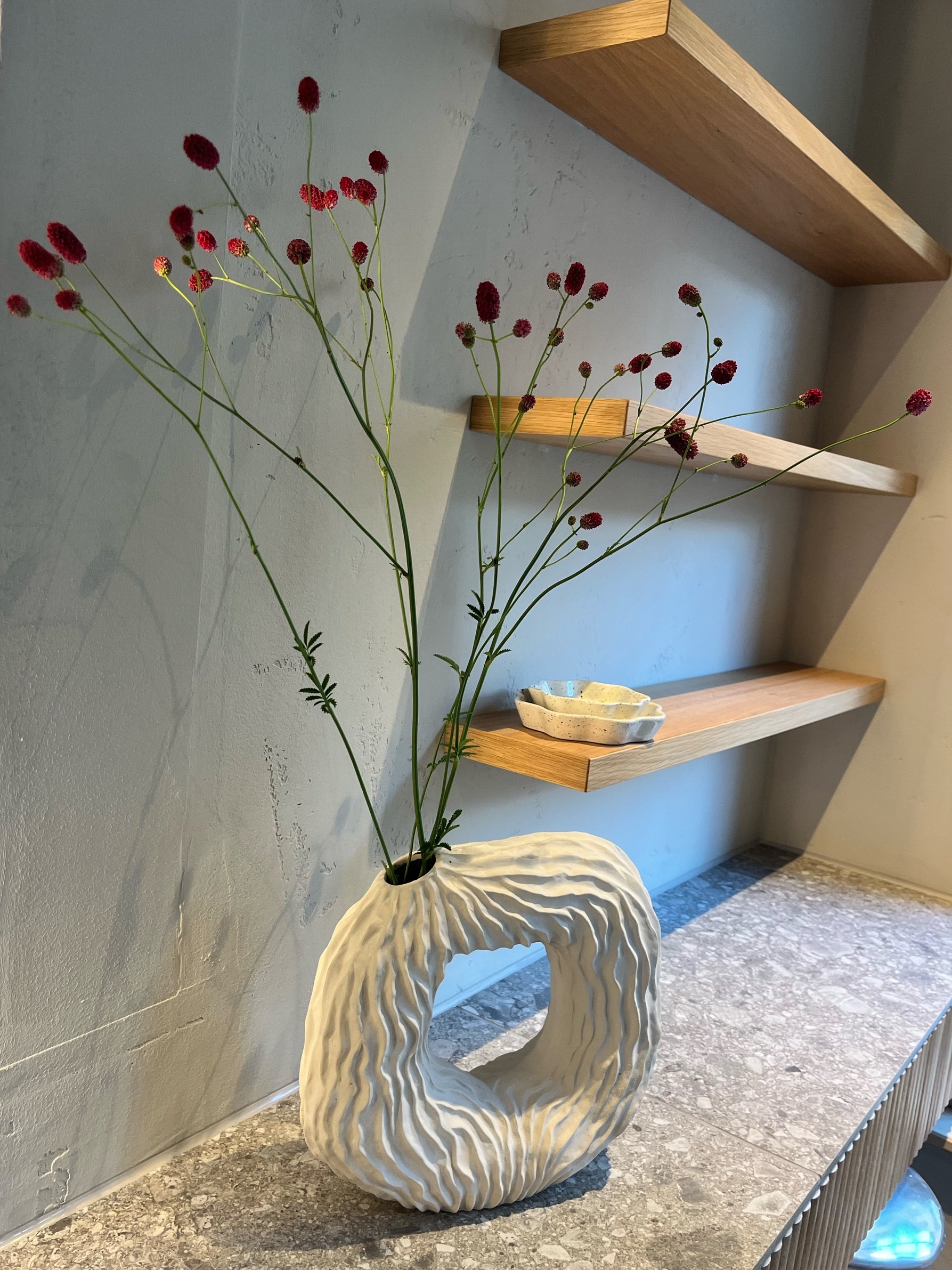 Kunstvolle weiße Vase mit geripptem Design, gefüllt mit langen Stielen und roten Blumen, auf einer Steinplatte, im Hintergrund Holzregale an einer hellen Wand.