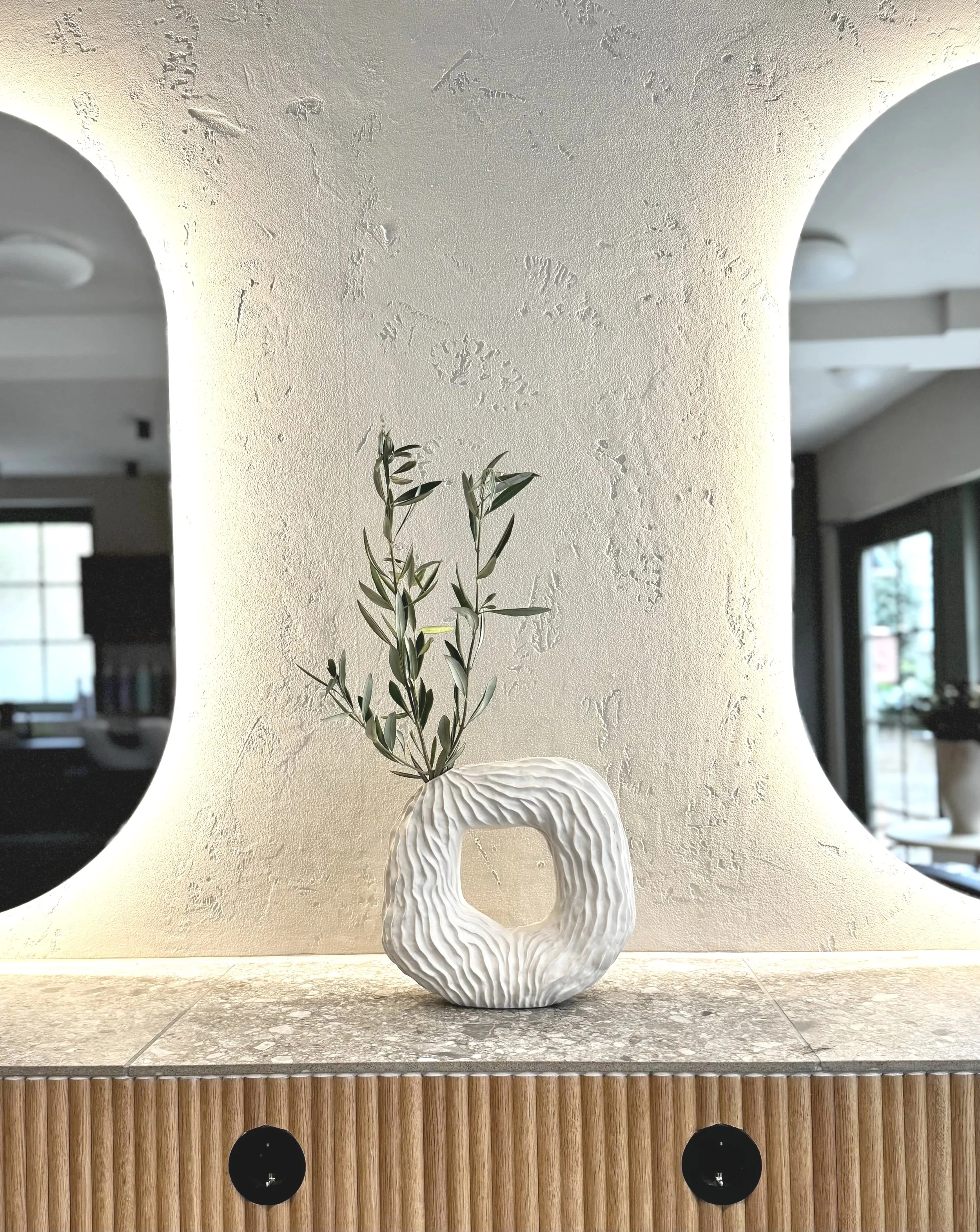 Weiße, strukturierte Keramikvase mit grünen Zweigen auf einer Holzoberfläche vor einer hellen Wand mit ovalen Ausschnitten.
