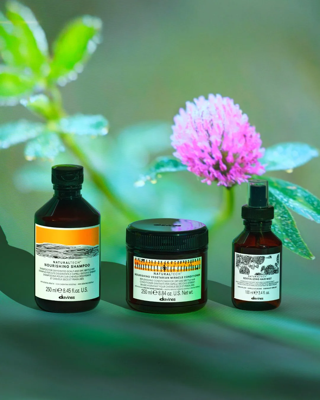 Drei Pflegeprodukte von Davines vor einem Hintergrund mit einer pinken Blume und grünen Blättern, einschließlich Nourishing Shampoo, Nourishing Vegetarian Miracle Conditioner und Revitalizing Hair Mist. Der Salon Esslingen, Friseursalon, Balayage