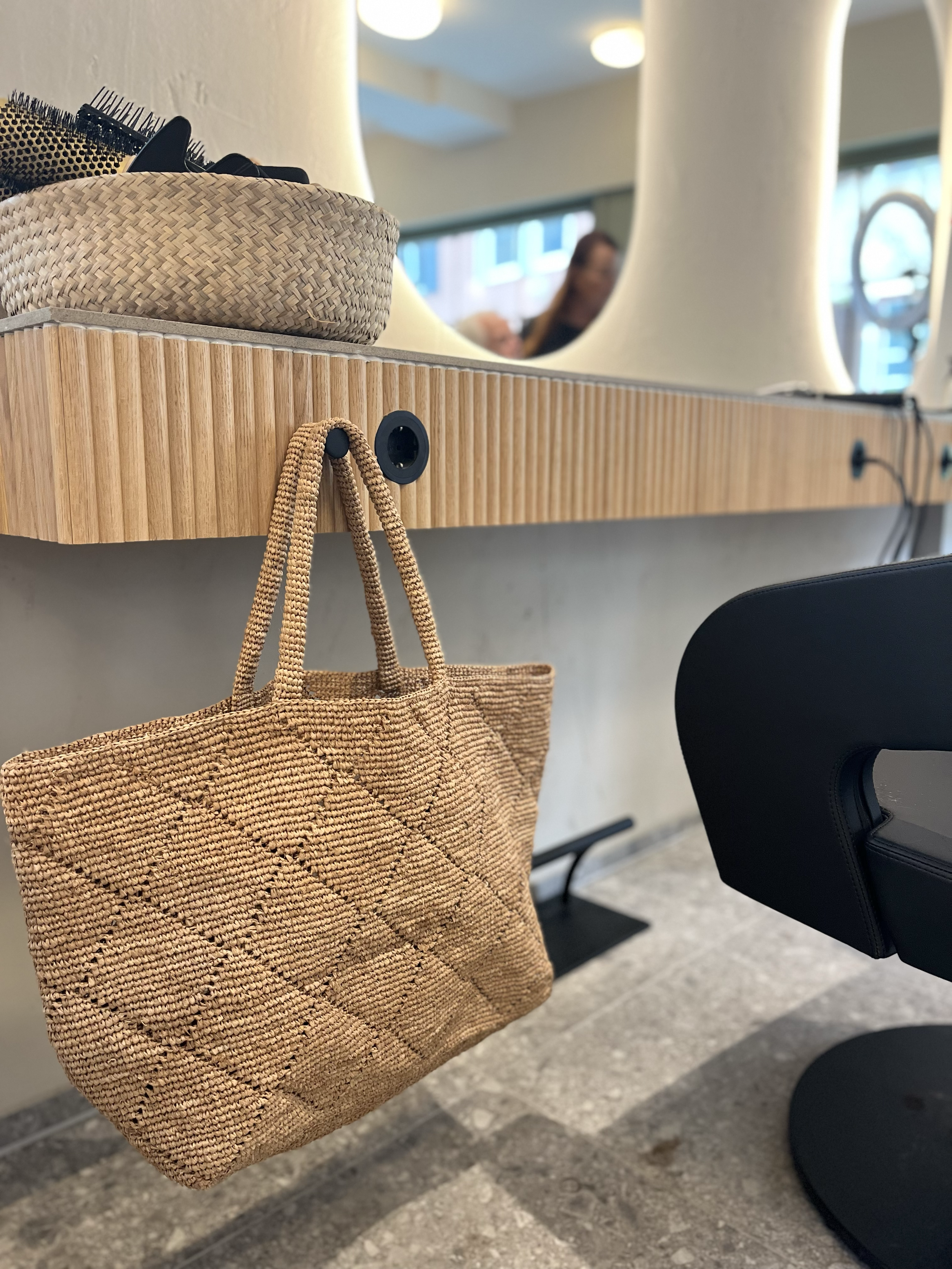 Ein Korbtasche hängt an einem Haken an einem Frisiertisch mit Spiegel, daneben sind Pinsel und andere Frisierutensilien in einer Schale. Im Hintergrund sind Menschen im Salon sichtbar.