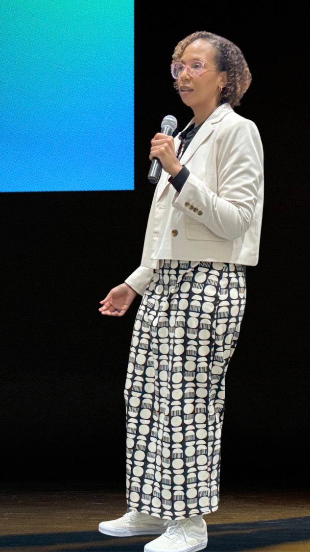 Woman standing on stage, holding a microphone, wearing glasses, a white blazer, patterned pants, and sneakers, speaking or presenting.
