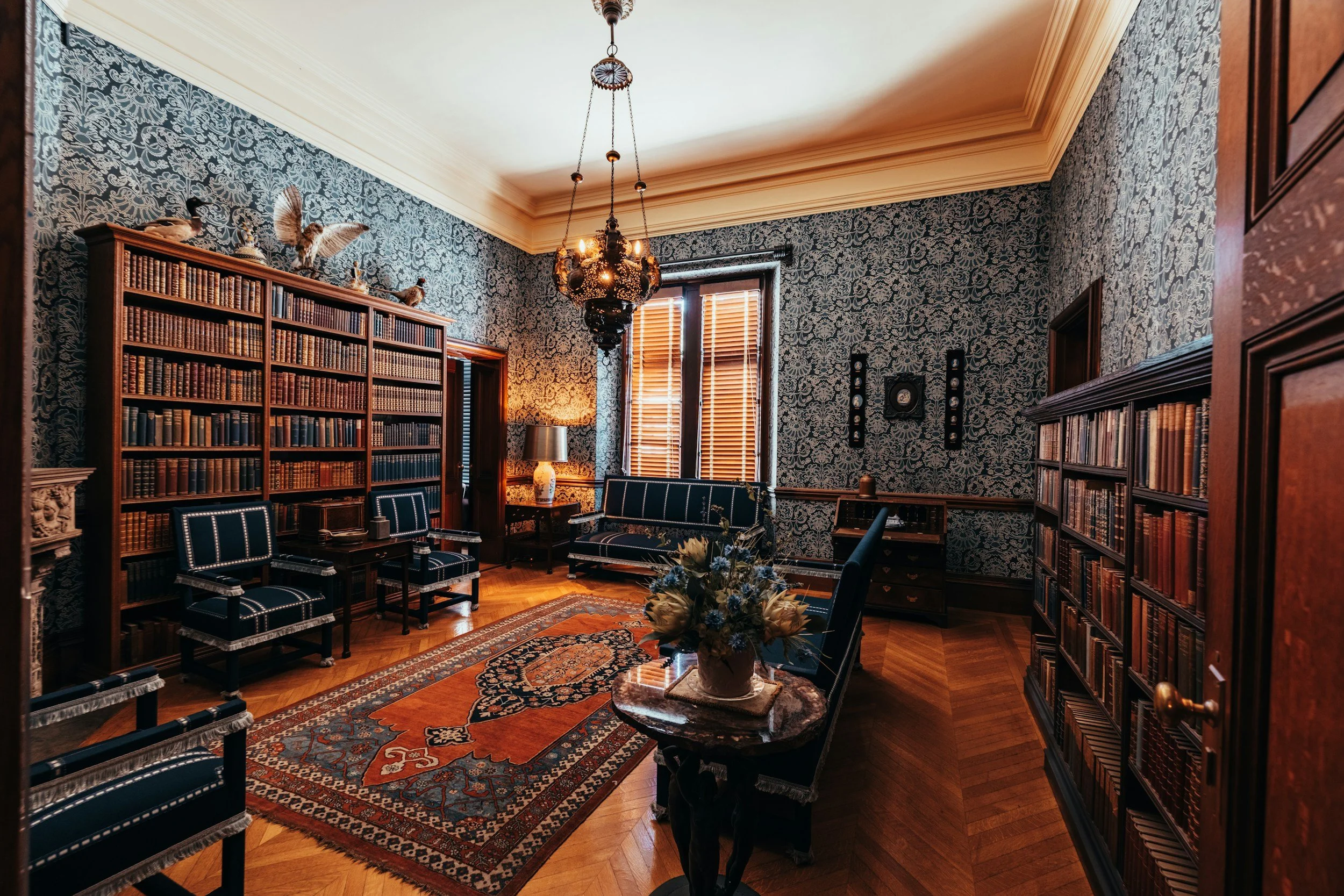 A vintage-style living room with dark wood bookshelves, ornate wallpaper, chandelier, and elegant seating.