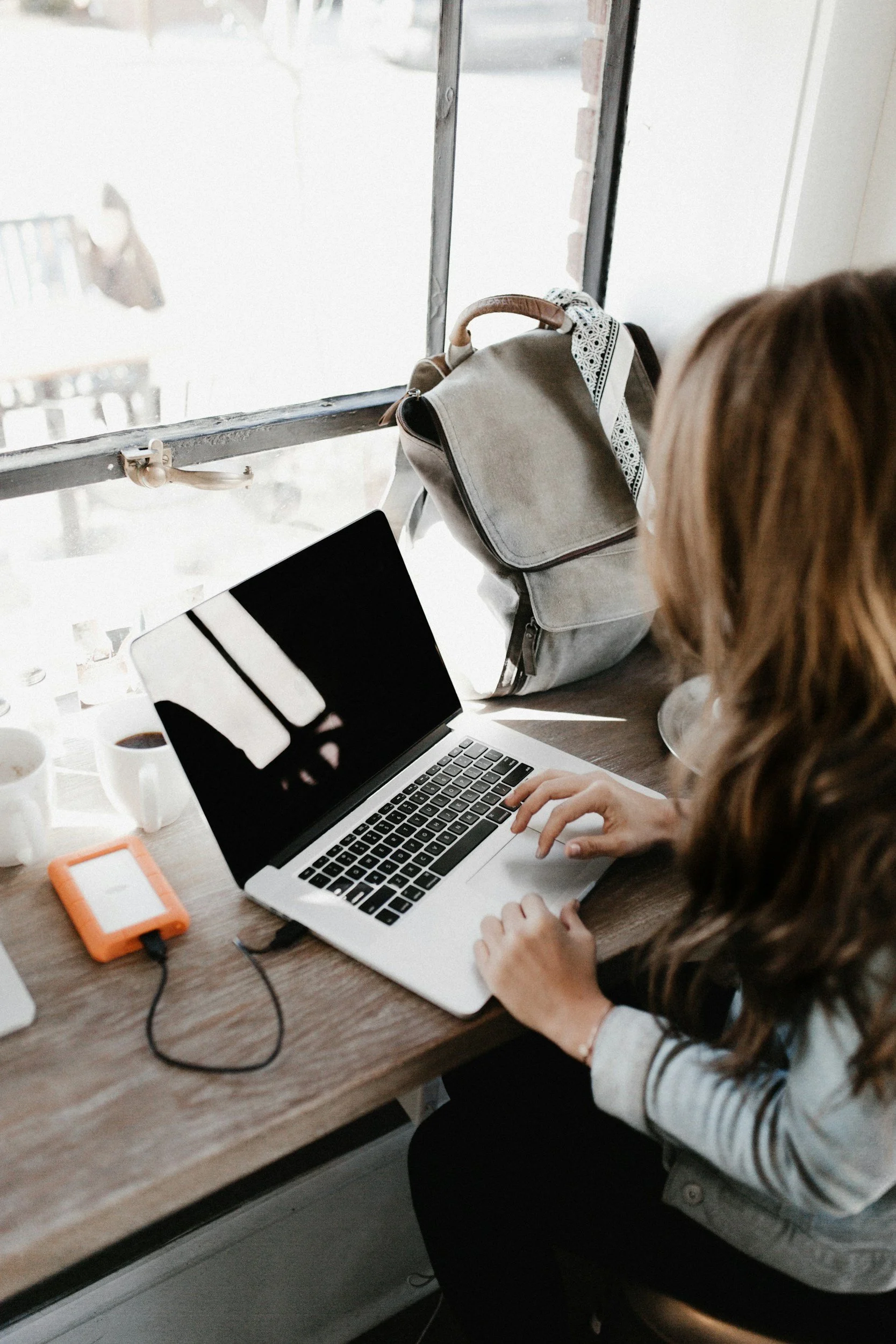 A woman working on a laptop at a wooden table, with a backpack and coffee cups nearby in a sunlit cafe.