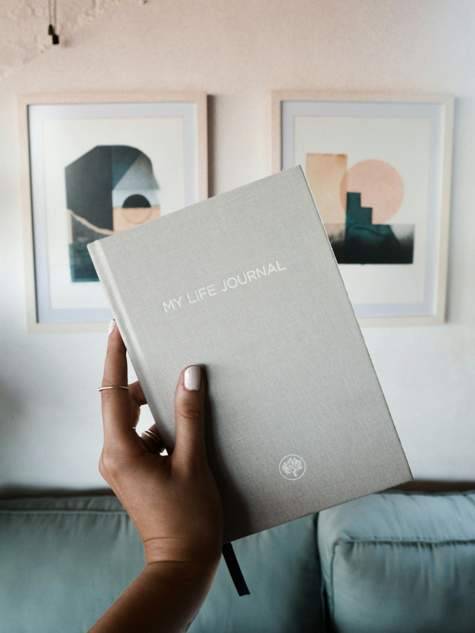 Person holding a grey journal titled "My Life Journal" in front of abstract art prints on a white wall.