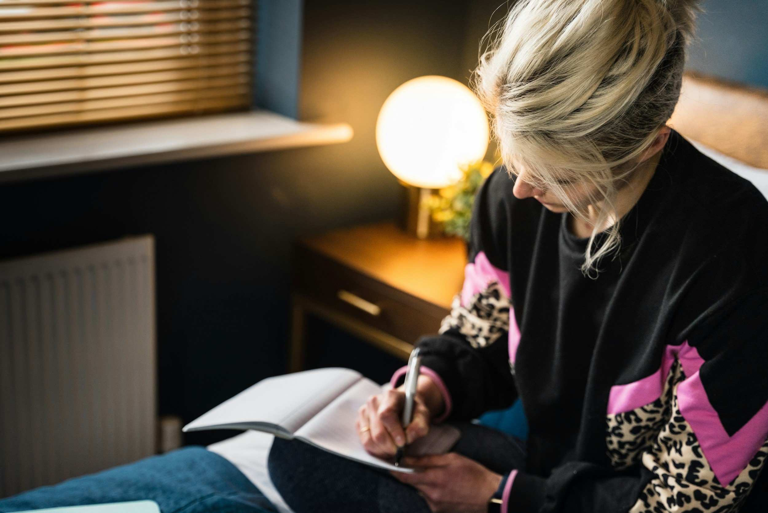 A woman with blonde hair sitting on a bed, writing in a notebook with a pen. There is a bedside table with a lamp turned on and a small plant behind her. The room is dimly lit.