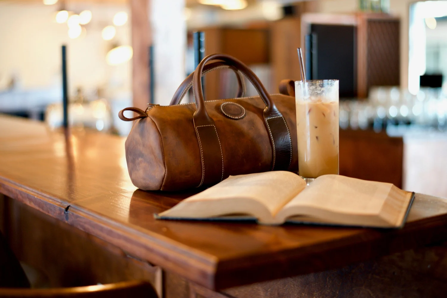 A quiet moment in a real day — an open book and coffee beside her leather bag on a café counter