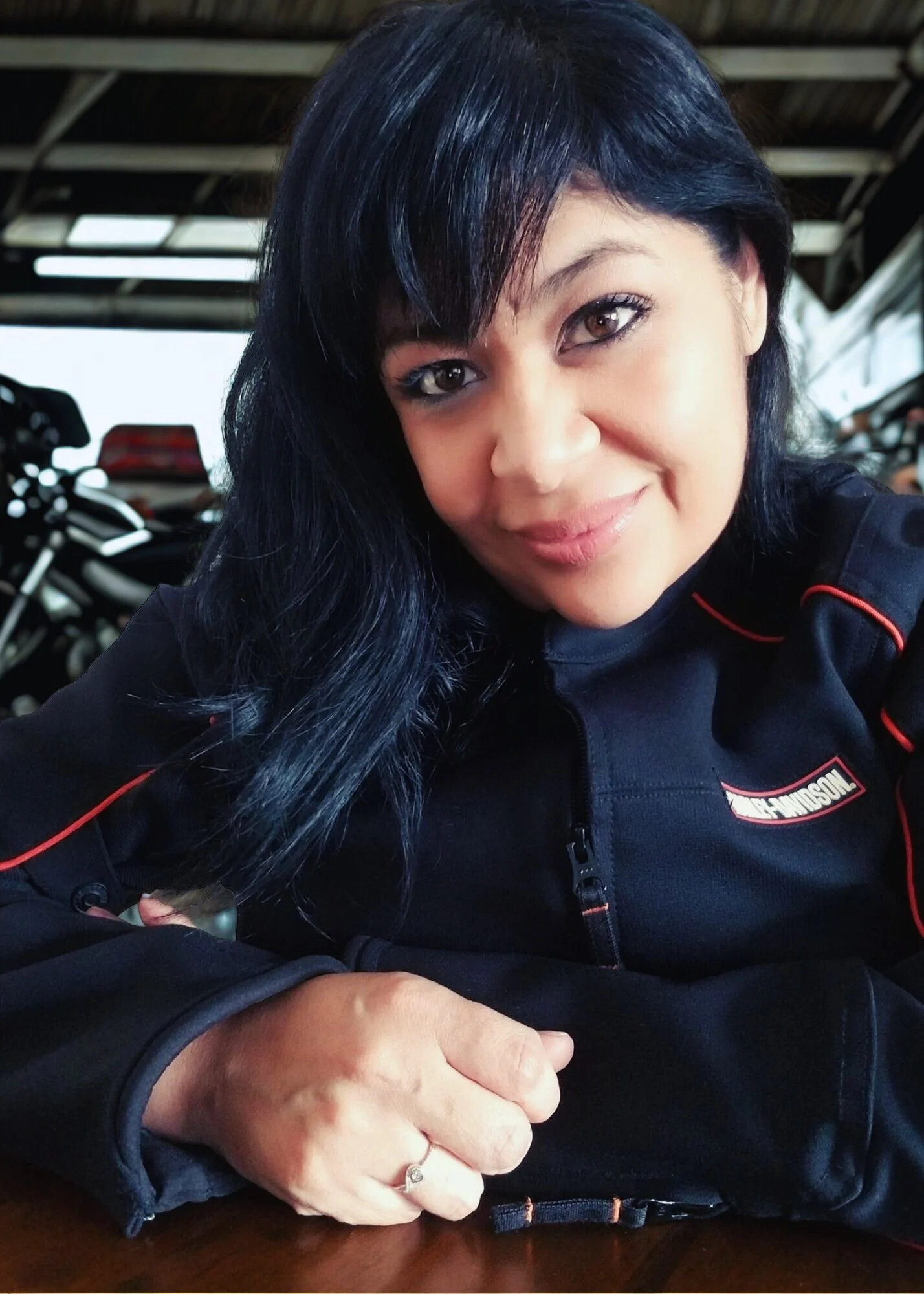 Close-up of Sandy Brooke wearing a black and red Harley-Davidson jacket, smiling and sitting at a table with motorcycles visible in the background.
