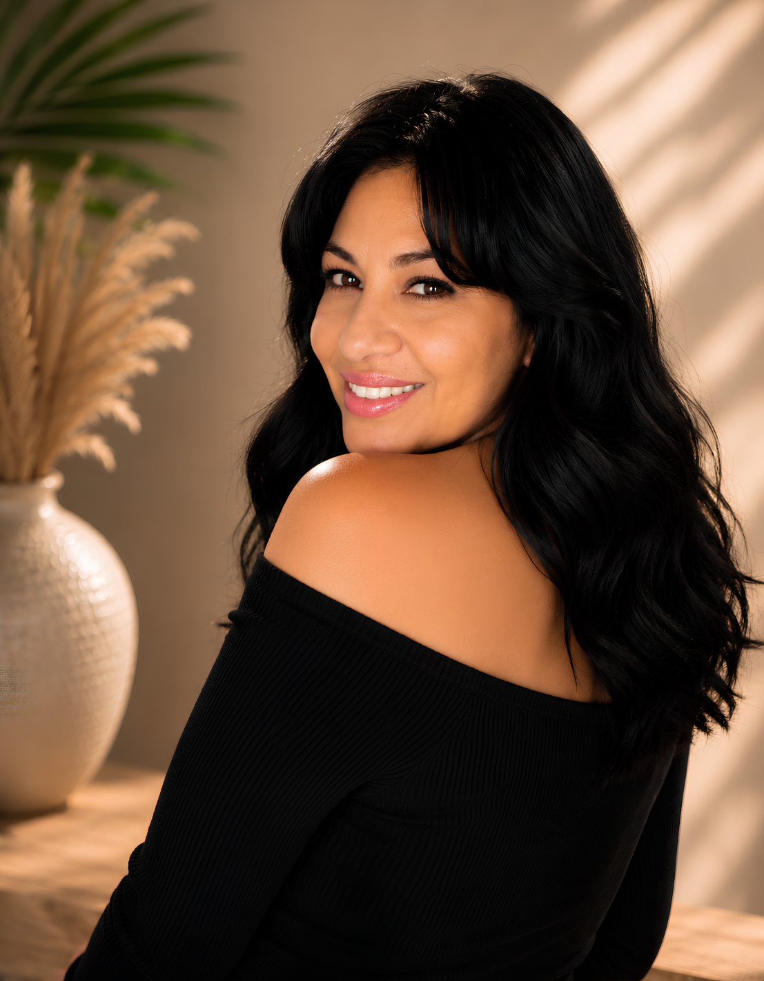 Sandy Brooke with long black hair smiling and looking over her shoulder, wearing a black off-the-shoulder top, with a background of a beige wall and a vase with dried plants.