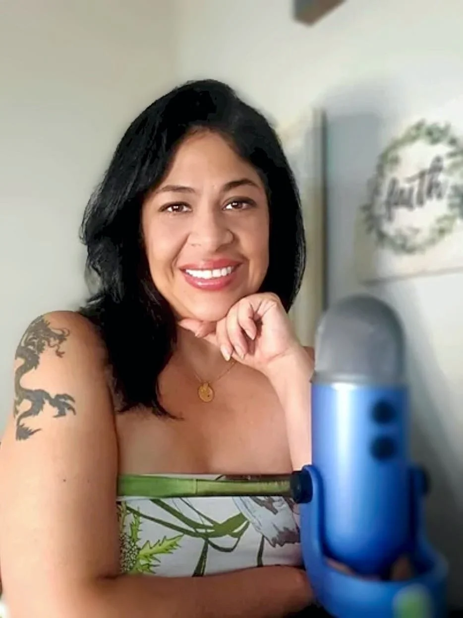 Sandy Brooke is smiling and sitting behind a blue microphone, wearing a strapless top with a green and white pattern.