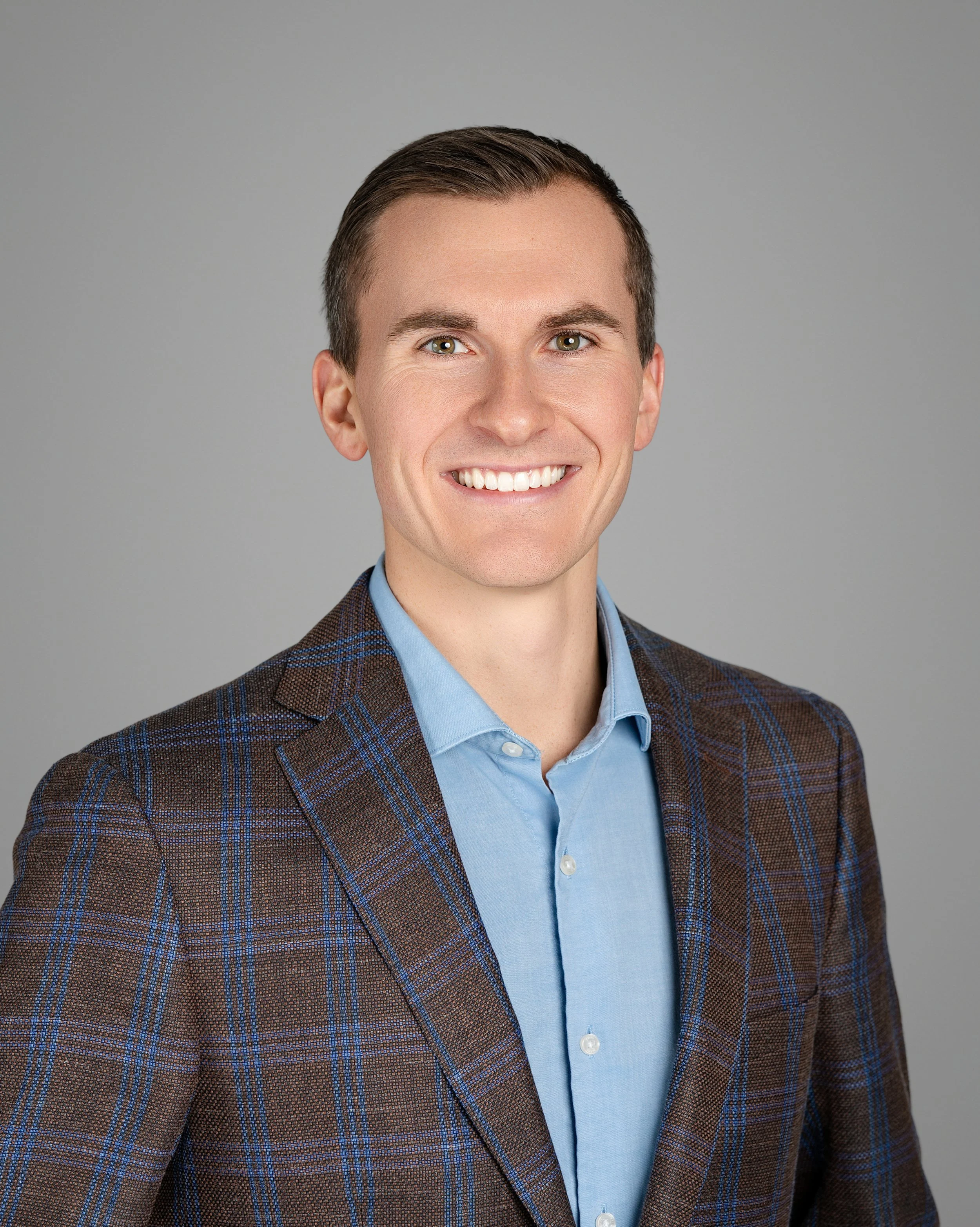 Headshot of a young man with short brown hair, dressed in a brown plaid blazer and light blue shirt, smiling against a plain gray background.