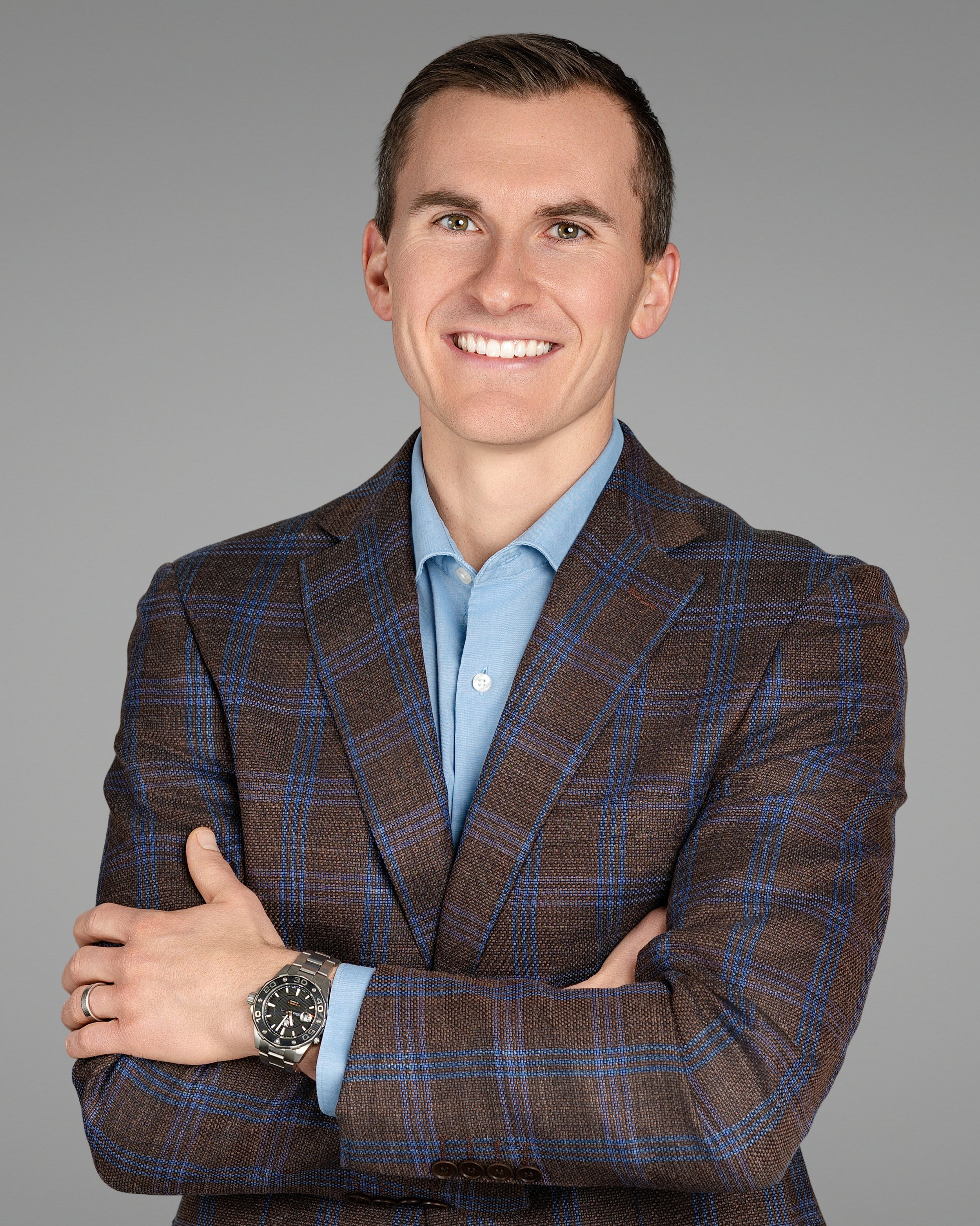 Headshot of a smiling man with short brown hair, wearing a brown plaid blazer and a light blue dress shirt, with crossed arms, against a plain gray background.