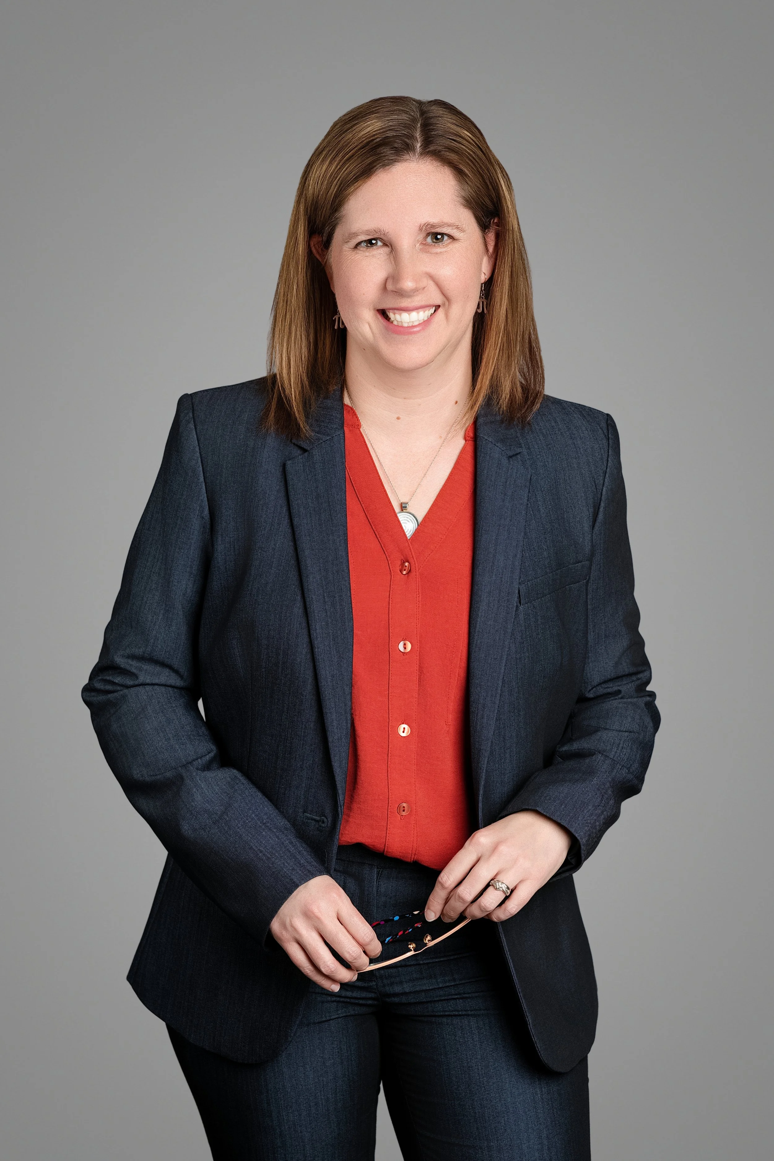 A woman with shoulder-length brown hair smiling, wearing a dark blazer over a red blouse, holding eyeglasses, against a plain gray background.