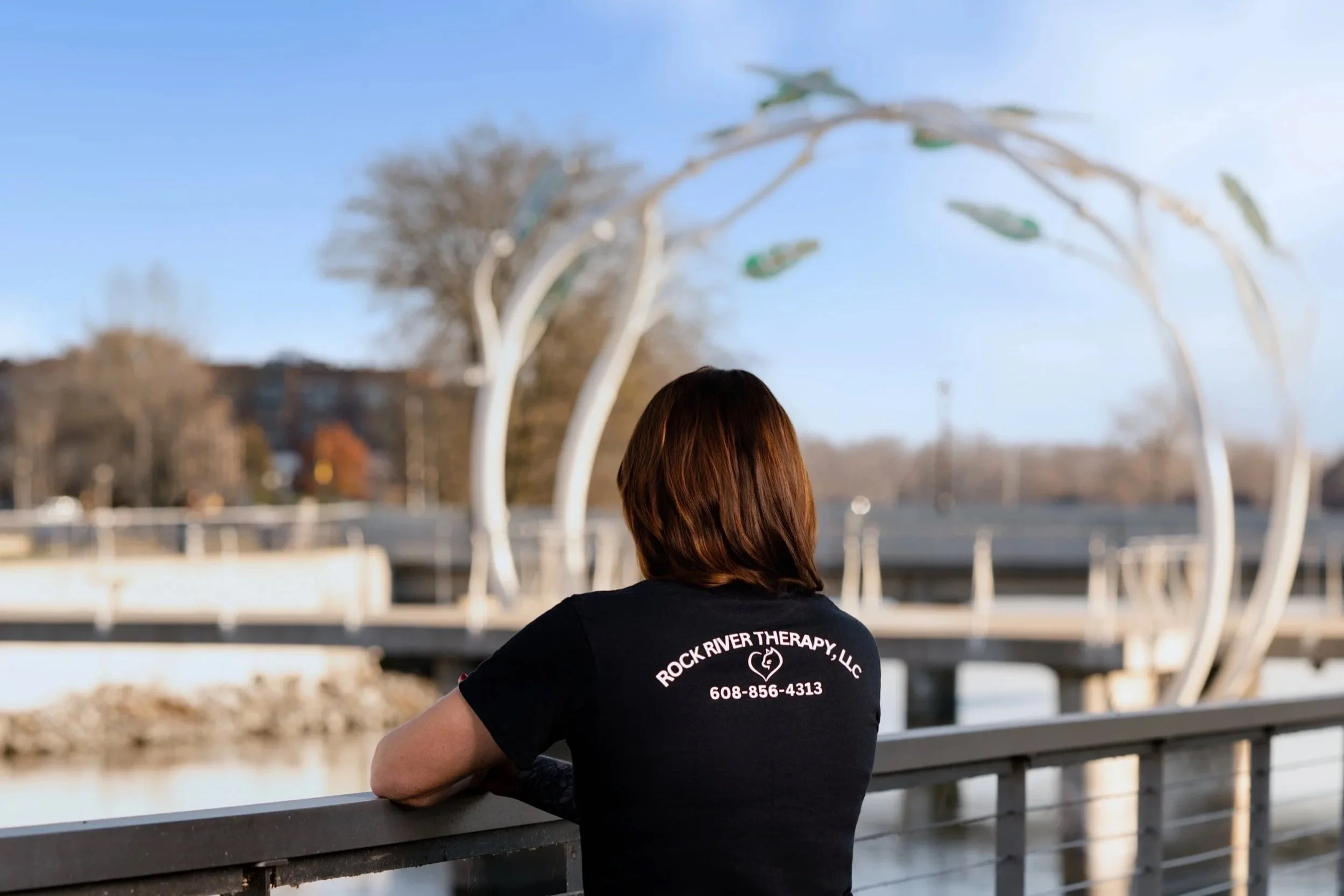 A woman with brown hair wearing a black T-shirt with 'Rock River Therapy, LLC' and a phone number on the back, leaning on a railing by a body of water, facing away from the camera. In the background there is a decorative white metal arch and trees.