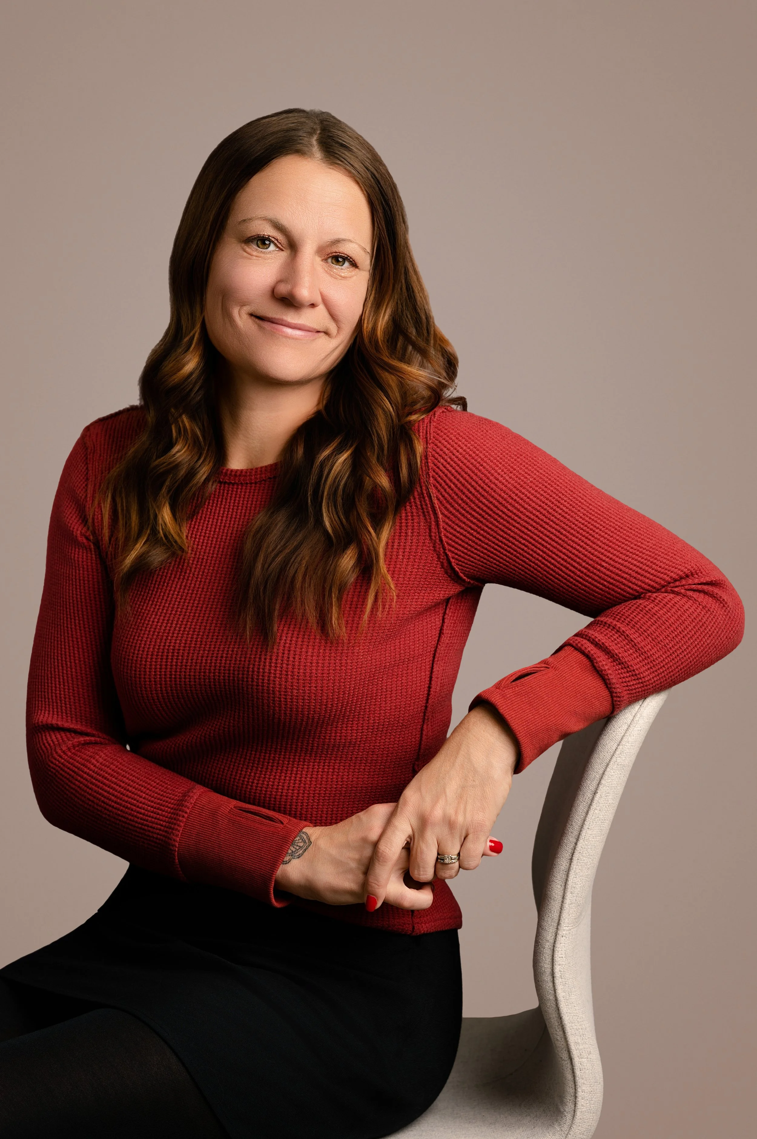 A woman with long brown hair wearing a red long-sleeve top and a black skirt, sitting on a white chair against a plain background.