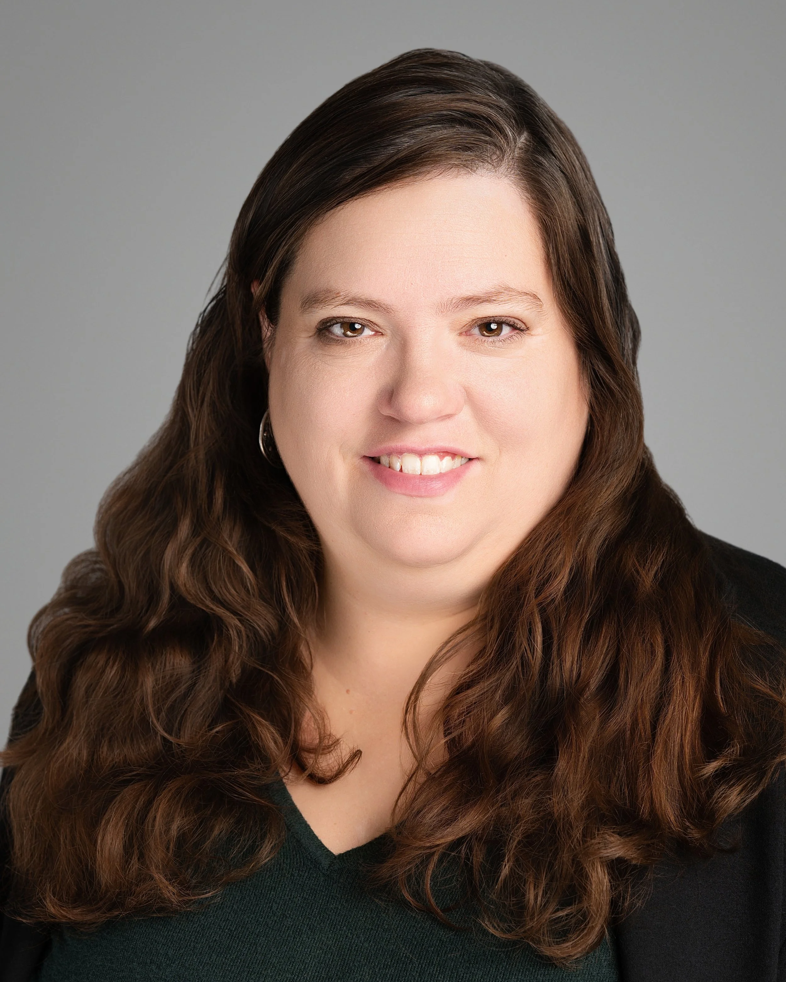 Professional headshot of a woman with long, wavy brown hair, wearing a black blazer and a dark green top, smiling against a gray background.