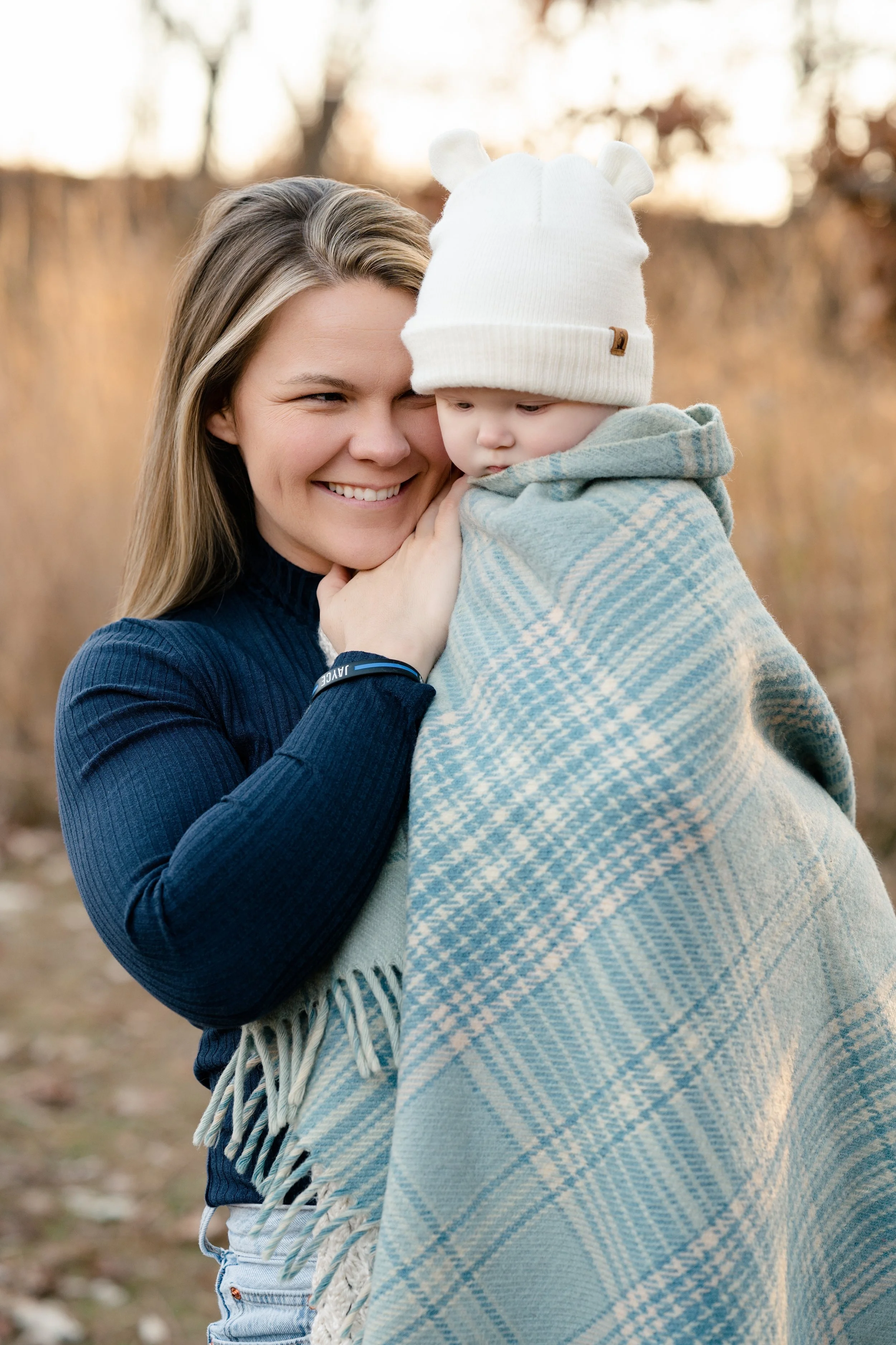 A woman holding a young child wrapped in a blue and white checkered blanket outdoors in a fall setting.