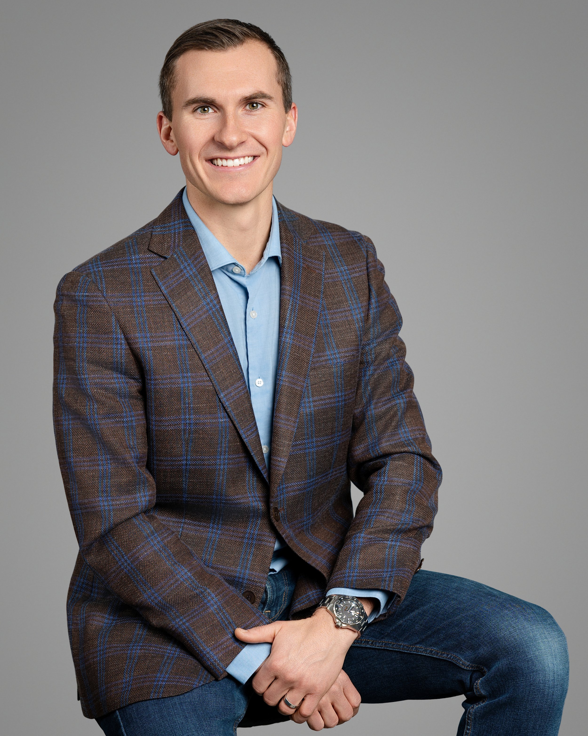 A young man with short hair, smiling, wearing a brown plaid blazer, light blue shirt, and jeans, seated against a gray background.