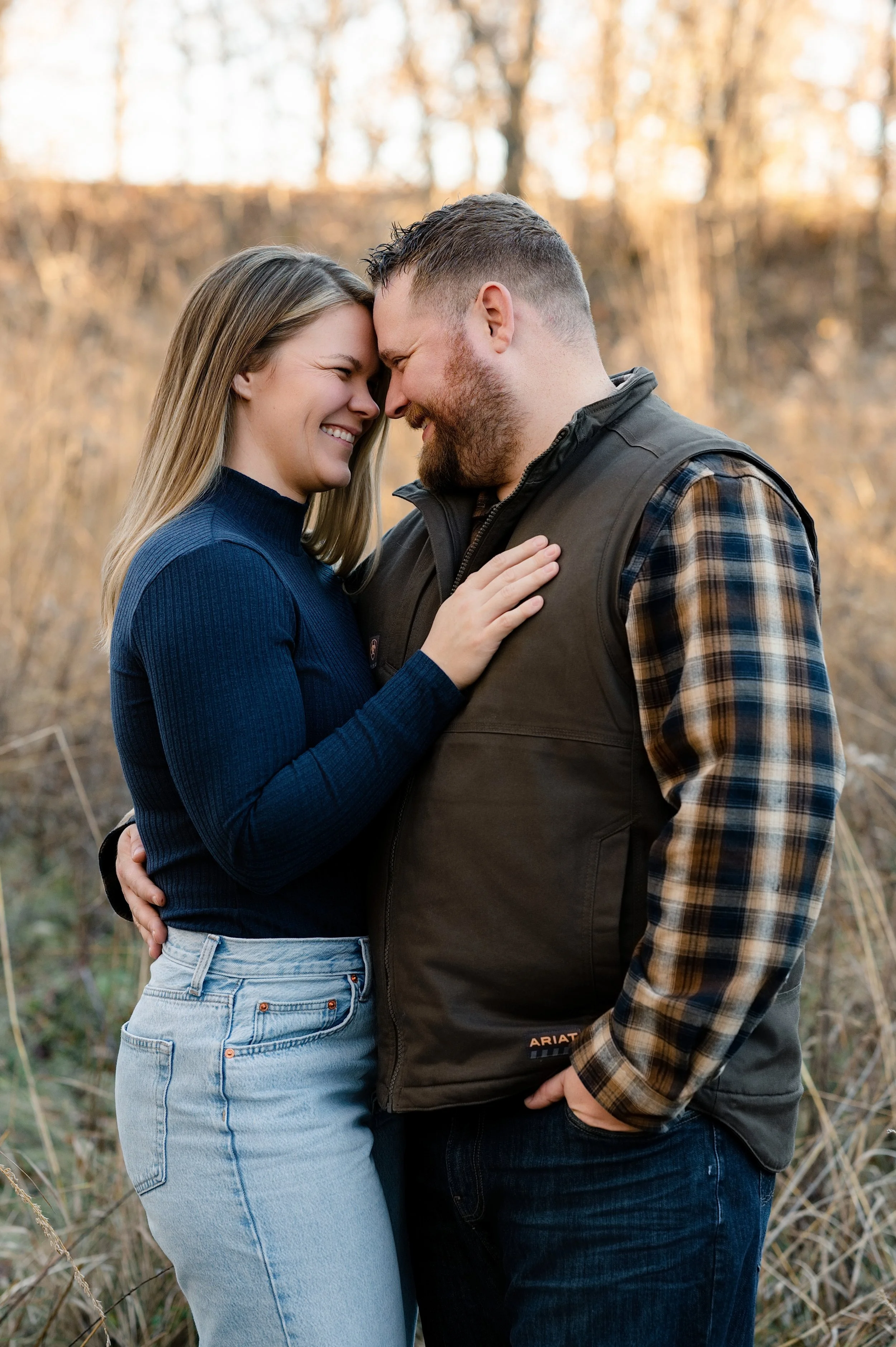 A happy couple standing close together outdoors, touching foreheads and smiling during sunset in a wooded area.