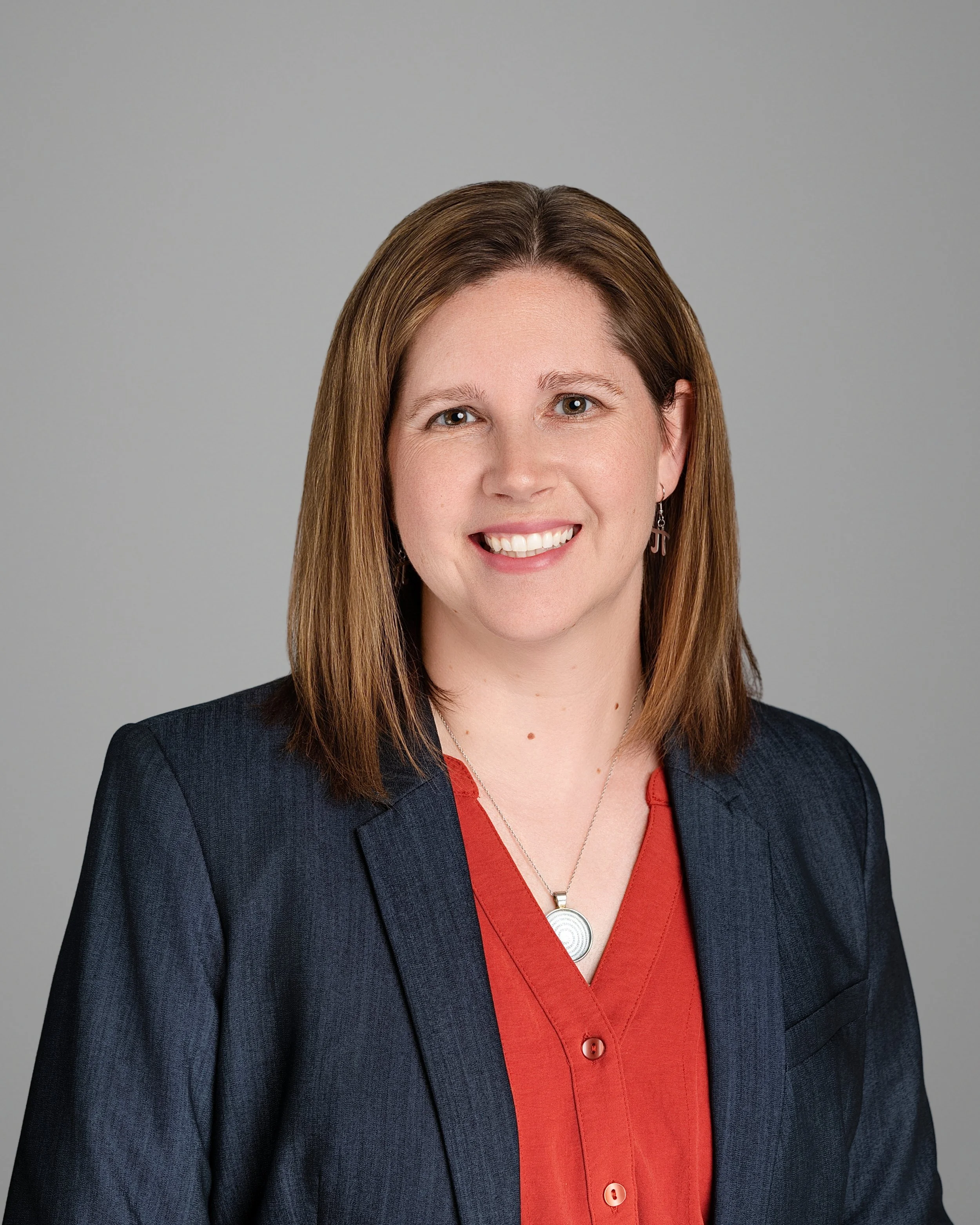 Portrait of a woman with shoulder-length brown hair wearing a black blazer and red blouse, smiling against a plain gray background.